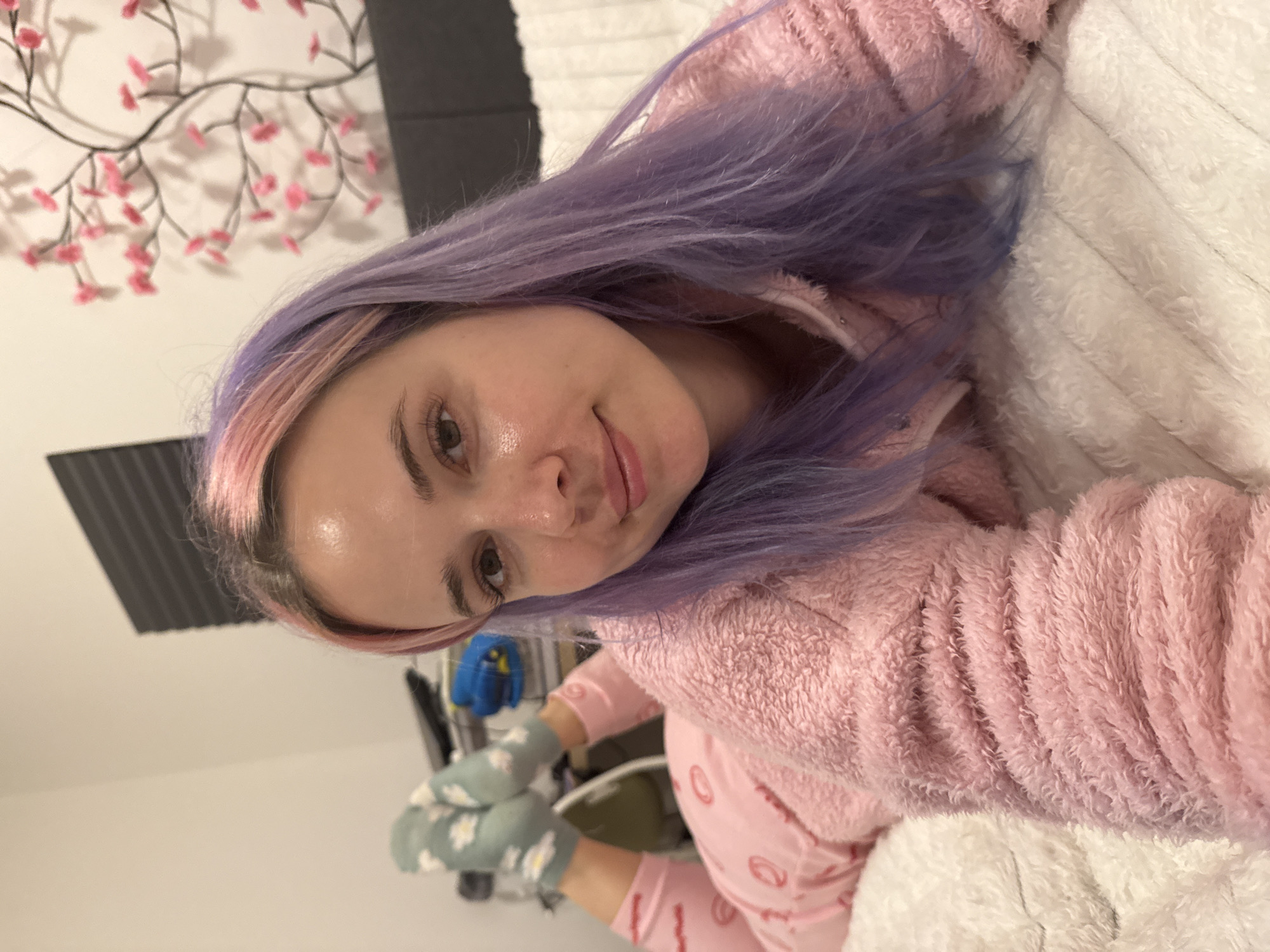 A female individual is depicted in a close-up, chest-up shot, lying prone on a bed within an interior space, identified as the City of Zagreb, Croatia. She faces the camera directly with a slight smile. Her long hair is dyed purple with pink highlights near the front. Her attire consists of a pink, fuzzy long-sleeved hooded top and pink pajama bottoms featuring a repeating red circular pattern. Her legs are bent at the knees, with her feet, clad in light green socks adorned with white daisy patterns, visible in the background. She rests on white, textured bedding. The background reveals a dark headboard, a white wall, and a decorative element resembling a dark tree branch with pink floral accents. In the upper left quadrant, a window with dark horizontal blinds is partially visible, alongside blurred objects on a surface, including a prominent blue item. The scene is brightly illuminated by artificial lighting.