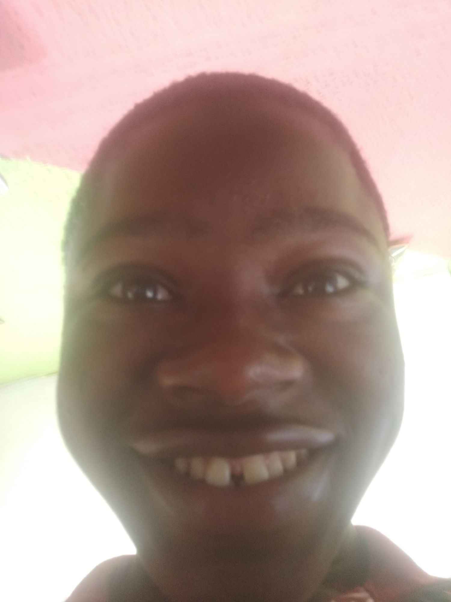A close-up, selfie-style photograph captures a smiling person with dark skin. The person has short, dark hair, and their face fills the frame. Their eyes are open and appear to be focused on the camera. The person's teeth are visible, and a gap between the upper front teeth is noticeable.
The background is somewhat indistinct, with what appears to be a blend of light pink, green, and white surfaces suggesting an indoor setting. The location in Shangisha, Nigeria, is speculative since there is no verifiable information.