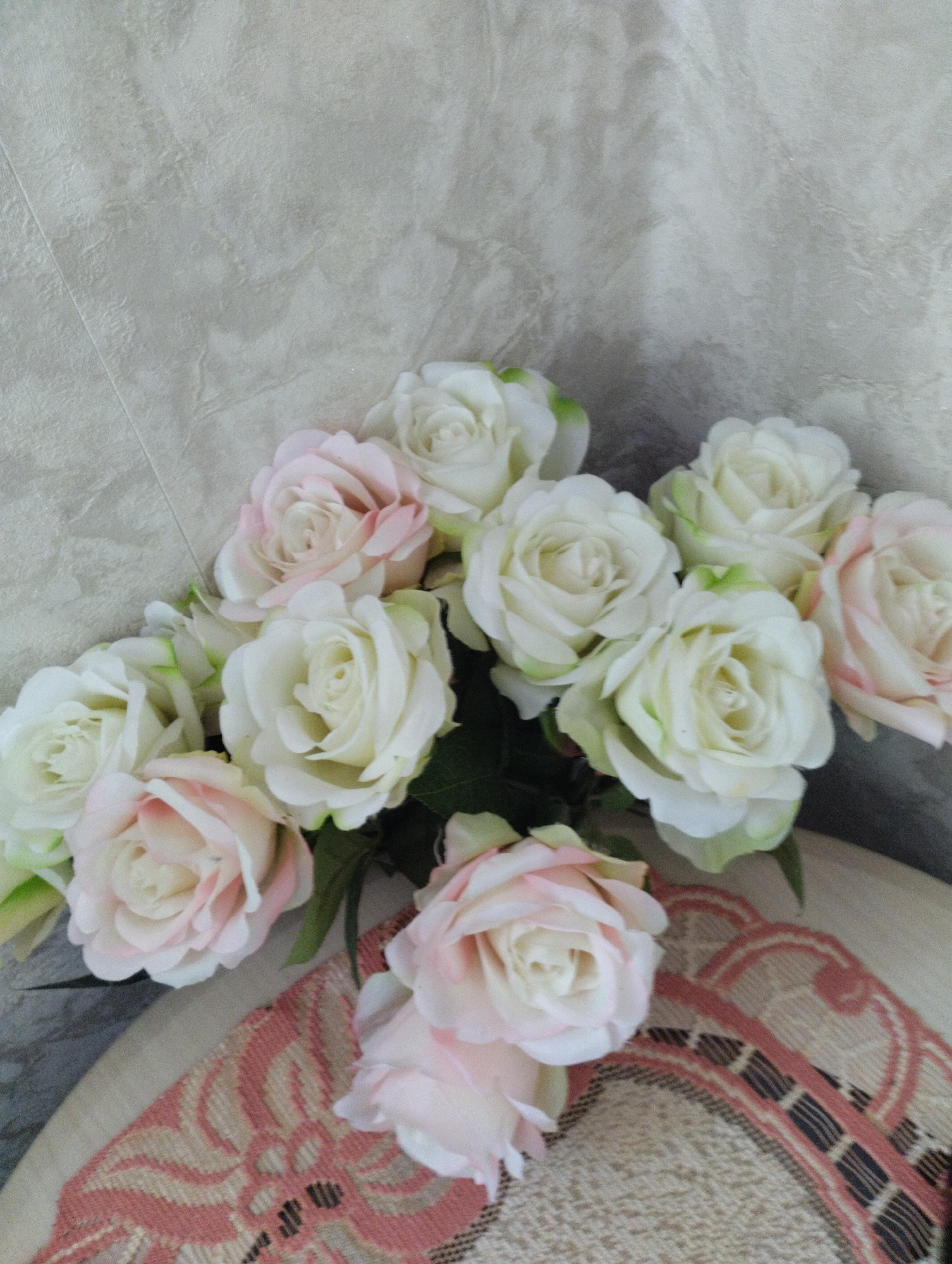 The image shows a bouquet of artificial roses, primarily white with some pink hues. The roses are arranged in a cluster, with some petals showing hints of green. They are placed on top of a textured, light-colored surface, which appears to be a textile with a woven pattern in shades of pink and beige. The background is a plain, textured wall or fabric in a light gray-beige color. No people, actions, or interactions are visible. The scene is static and appears to be an indoor setting. The location in Astravyets, Belarus, is not discernible from the visual elements of the image.