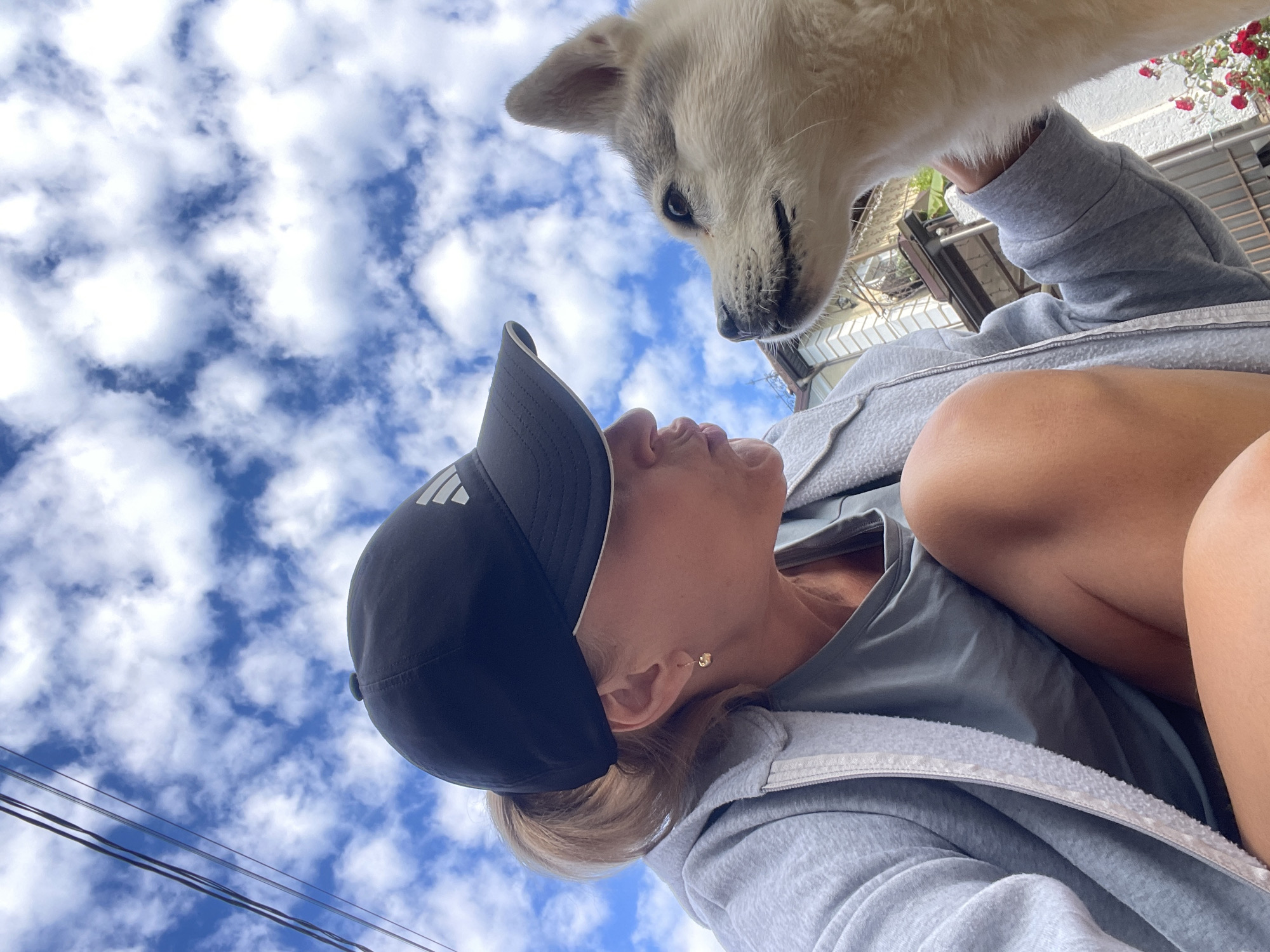 A low-angle shot shows a woman and a white dog. The woman is wearing a black baseball cap with the Adidas logo, a gray hoodie, and a gray shirt. She is looking up at the dog, who is also looking down at her. The background is a blue sky with white clouds. Part of a building with red flowers is visible at the top right. The location is possibly Baranovka, Russia, but without additional context, this cannot be confirmed.