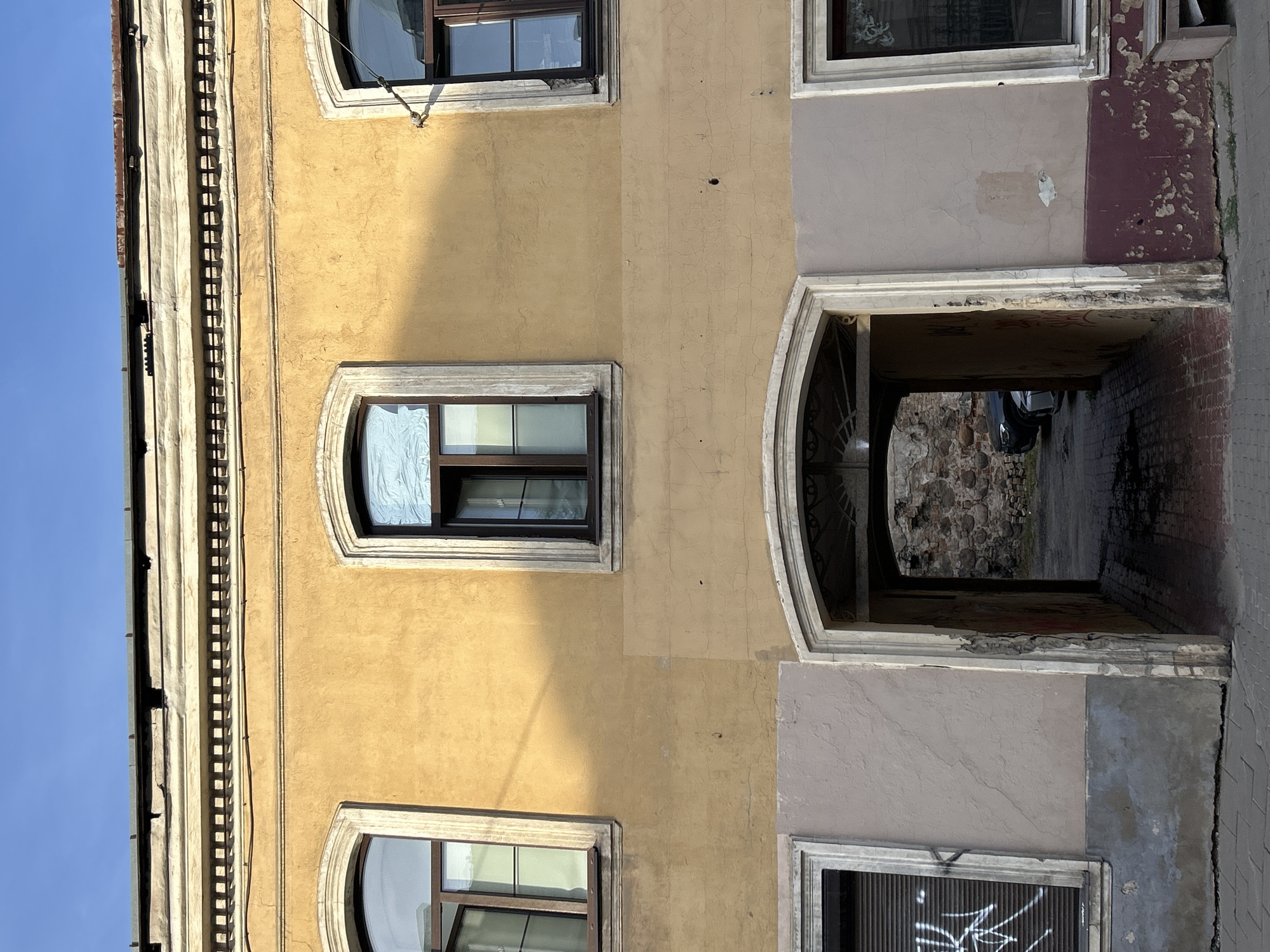 The image shows the facade of an old building with a courtyard entrance, located in Vilnius, Lithuania. The building is constructed from brick and stucco, painted in shades of yellow and beige. The upper part of the facade features a decorative cornice with a band of small, square openings. There are three arched windows on the upper floor, with two of them visible in the frame. The windows have wooden frames and appear to be partially open.
Below the upper windows, there is a large arched entryway that leads into a courtyard. The entryway is framed by a stone arch and appears to be dimly lit. Inside the courtyard, stone walls and a parked car are visible.
On the ground floor, to the left of the entryway, is a window with a closed metal shutter that has white graffiti on it. To the right of the entryway is another window with a dark frame. The lower part of the building shows signs of wear and peeling paint, with a maroon-colored section at the bottom. The ground in front of the building is paved with cobblestones. The sky above is clear and blue.