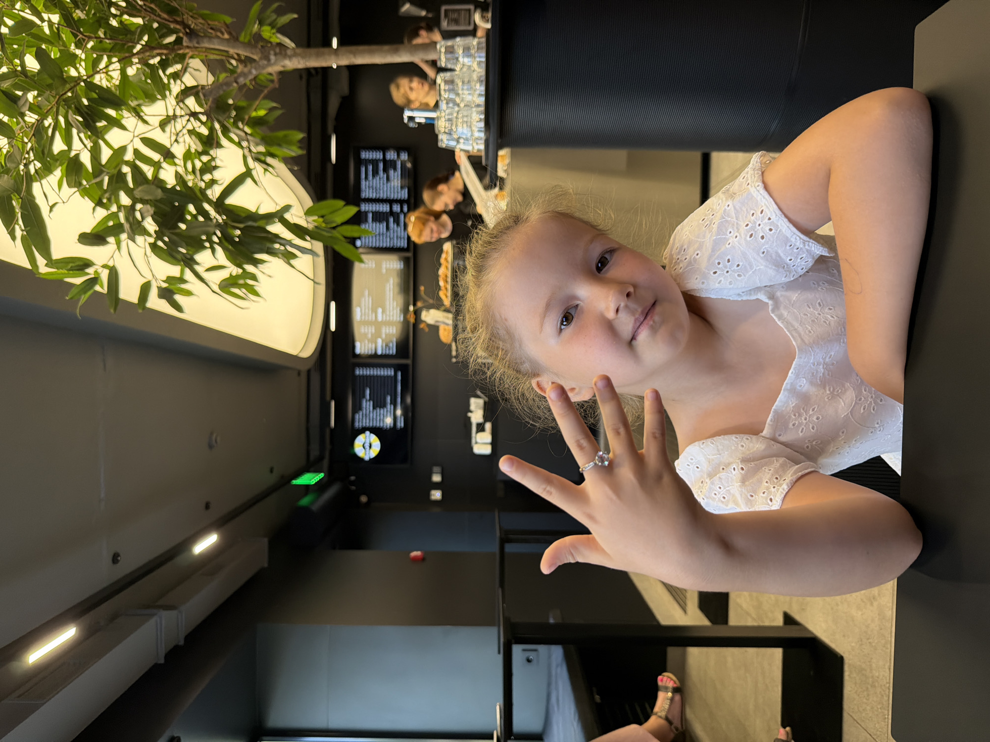 An outdoor portrait features a young girl sitting at a table in a modern cafe. The girl, with blonde hair and brown eyes, wears a white, embroidered top and is looking at the camera. She holds up her left hand, displaying a ring on her ring finger. In the background, a tree with green leaves is visible, along with a bright, diffused light source. The cafe environment includes other patrons, a counter with stacked glasses, and digital menu boards. The setting appears to be a contemporary establishment, possibly in Izhevsk, Russia, given the visual cues of the interior design and lighting.