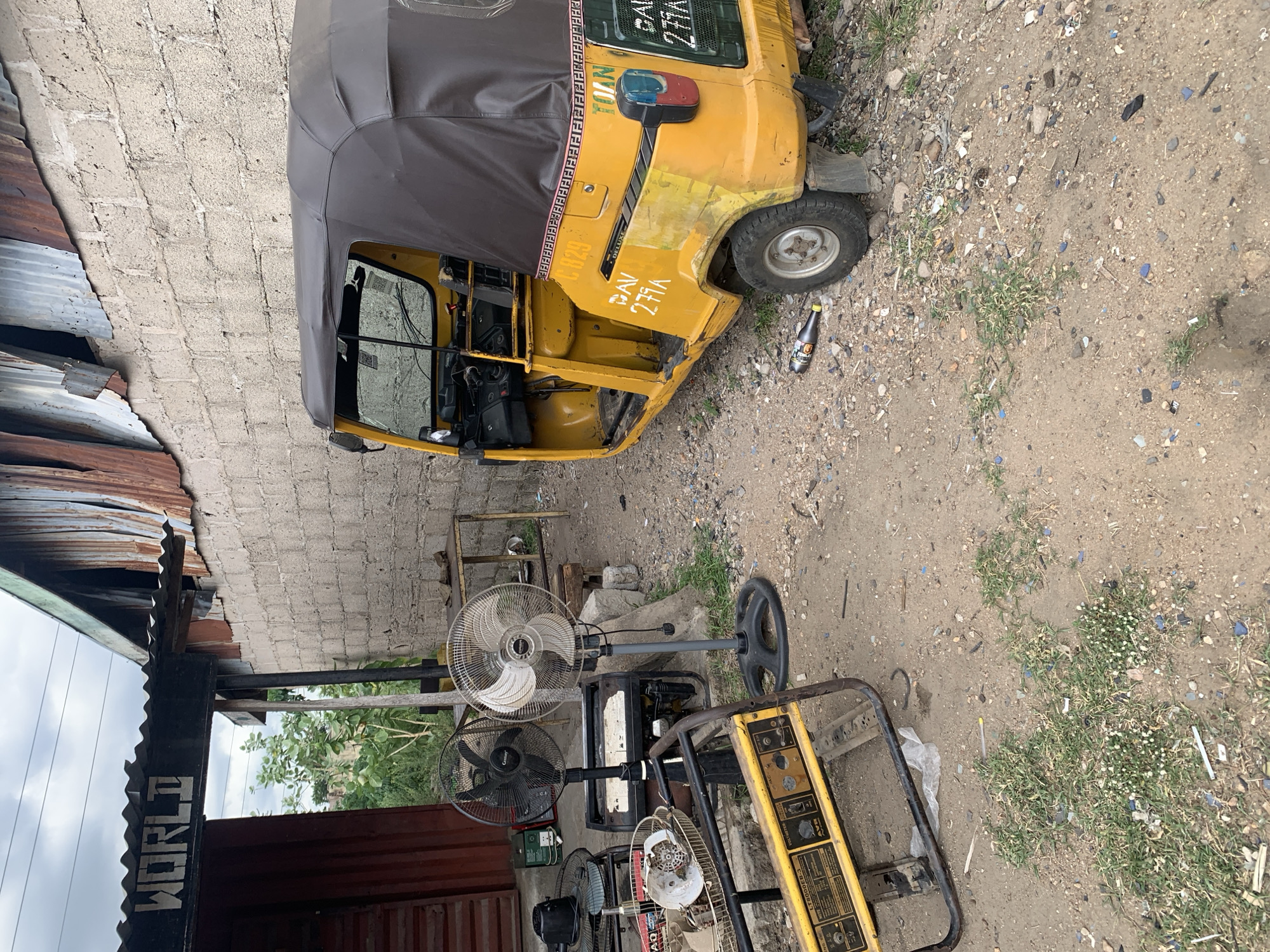The image depicts a scene in Adankolo, Nigeria. A yellow tricycle taxi (also known as a "Keke") is parked on dirt ground near a building. A bottle of drink lies beside one of the Keke's tires. A generator and several electric fans are nearby. A partially visible sign reads "WORLD". The ground is a mix of dirt, small rocks, and some vegetation. The building visible is constructed of cinder blocks and corrugated metal.