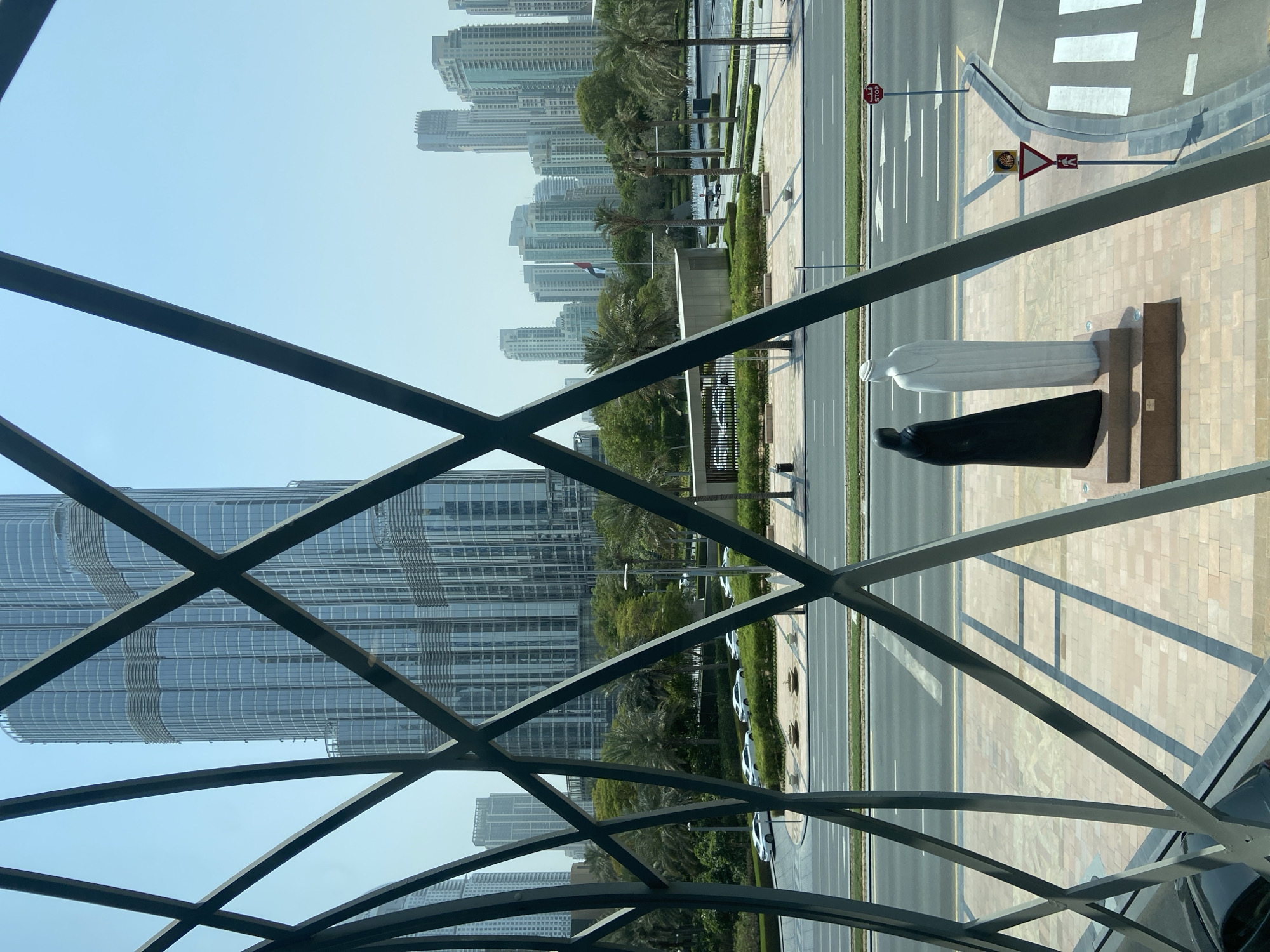 This image captures a view of the Burj Khalifa and surrounding cityscape in Dubai, United Arab Emirates, framed by an abstract metal structure. In the foreground, two statues stand on a patterned tiled plaza. One statue depicts a figure in a black abaya, and the other a figure in a white thobe. Behind them, a multi-lane road with white markings is visible, with several white cars driving on it. Beyond the road, a landscaped area with trees and palm trees separates the road from a modern building complex. Further in the background, the towering Burj Khalifa dominates the skyline, with other contemporary skyscrapers also present. The sky is clear and blue, suggesting a sunny day. A flag of the United Arab Emirates is visible on a pole in the distance. The overall scene depicts an urban environment with a blend of modern architecture, green spaces, and cultural elements.