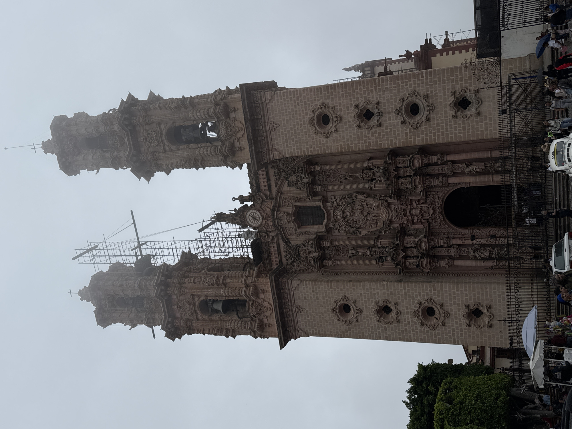 The image shows the Church of Santa Prisca in Taxco, Mexico, on a cloudy day. The church's ornate Baroque facade is visible, with two bell towers. Scaffolding is erected around the top of the left tower, indicating ongoing maintenance or restoration work. A large clock face is visible on the main body of the church, between the towers.
In the foreground, a black metal fence encloses the church grounds. Beyond the fence, there are several people walking and standing, some holding umbrellas. There are also market stalls and a white Volkswagen Beetle with the number "102" on its roof, suggesting a public plaza. Trees with lush green foliage are visible to the left of the church. The overall atmosphere is somber due to the overcast sky.