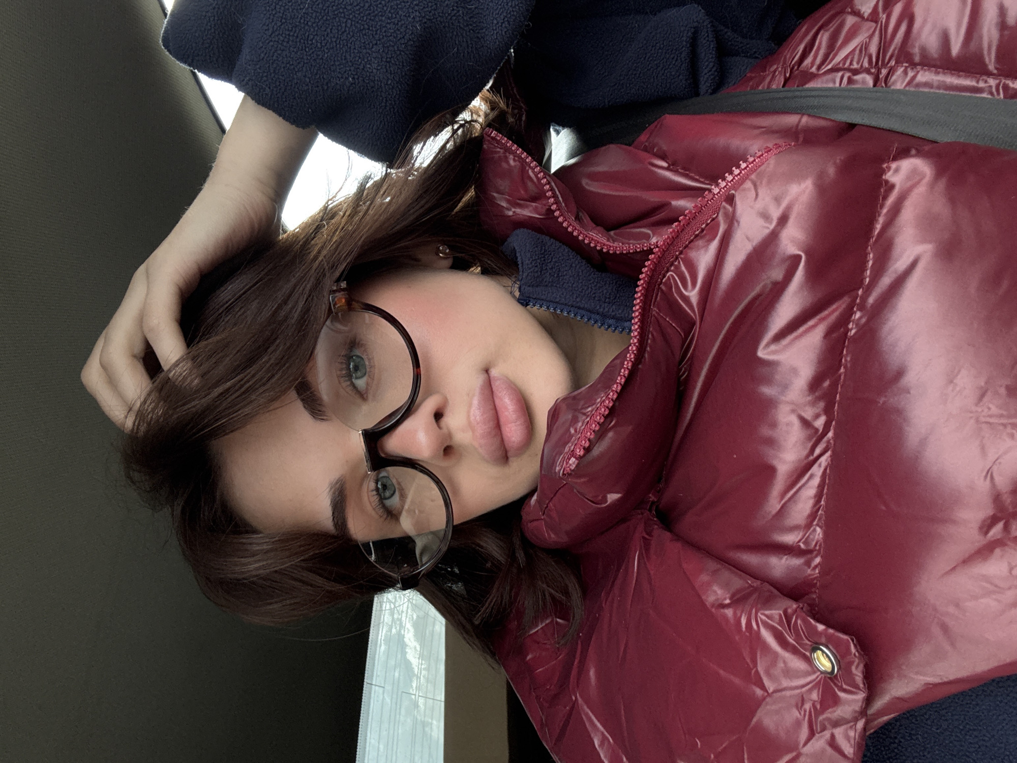 A close-up, slightly tilted portrait features a young woman looking directly at the camera. She has light skin, dark brown hair, prominent eyebrows, and light blue/green eyes. She wears round, tortoise-shell framed glasses and has a neutral to slightly pensive expression, with full lips. A subtle blush is visible on her cheeks.
She is dressed in a dark blue fleece top or jacket, layered under a shiny, puffy, dark red (burgundy) puffer jacket with a matching colored zipper visible down the front. A dark strap, potentially a seatbelt or a bag strap, crosses her chest. Her right hand is raised, with fingers gently touching her hair near the crown of her head. A small gold earring can be seen in her right ear.
The background is largely out of focus. To the left, there is a dark, solid surface. To the right and behind her, a lighter, indistinct area suggests a window or a bright light source. A faint reflection, possibly of a building or window blinds, is visible in the lower left portion of the background. A small part of a white face mask is also visible in one of the detailed crops, resting near the bottom right. The presence of a puffer jacket suggests a cool climate, aligning with the location context of Perm, Russia. The setting appears to be an interior space, possibly a vehicle or a room with a window. No text is visible in the image.