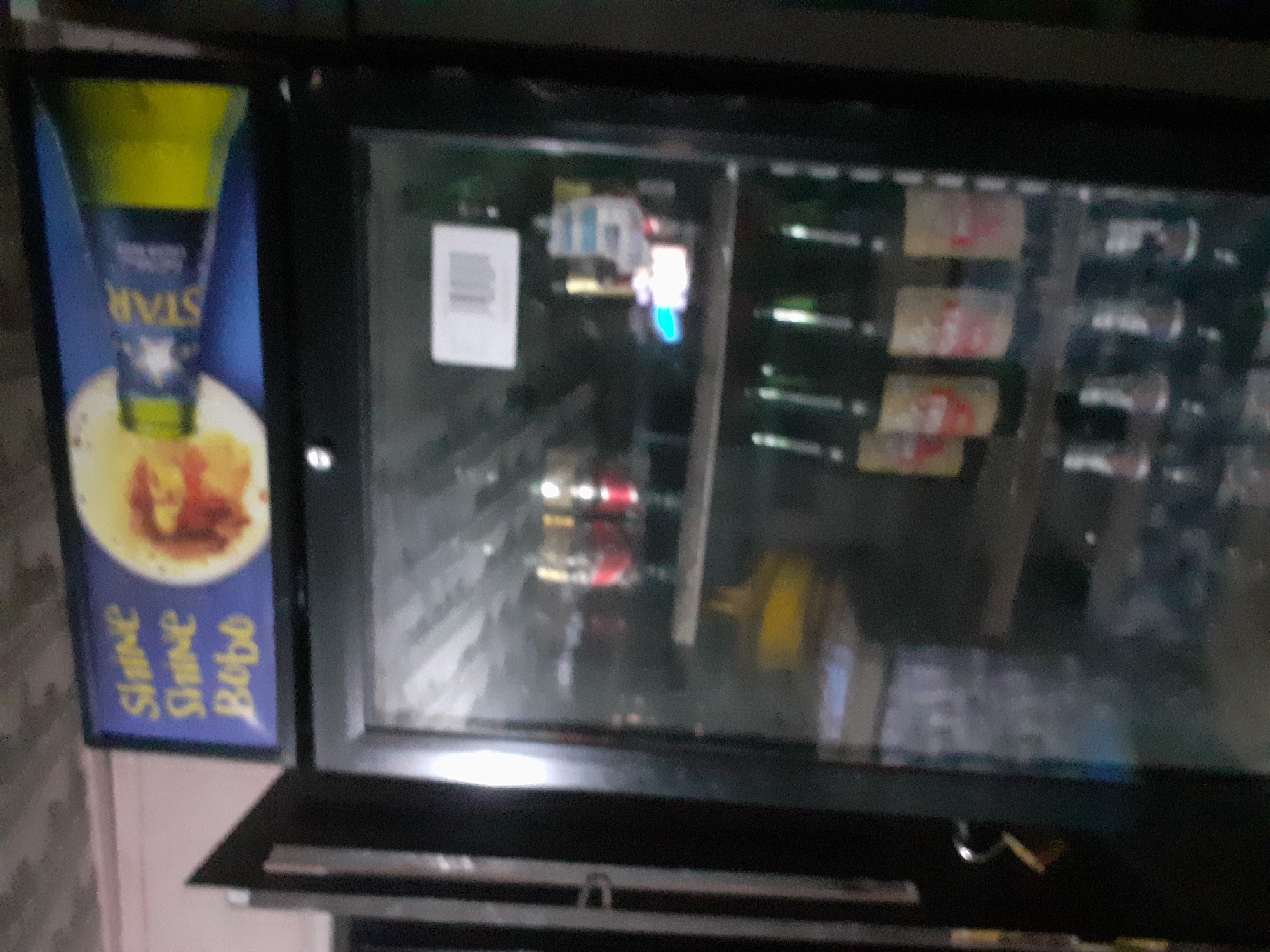 The image shows a blurry view of a commercial beverage cooler in Jalingo, Nigeria. The cooler has a glass door, revealing rows of bottled drinks. At the top of the cooler, a sign is visible with yellow text on a blue background, which reads "SHINE SHINE BOBO" and features an image of a bottle of Star beer. Inside the cooler, the drinks appear to be mostly bottled beer and possibly some soft drinks, with labels that are largely unreadable due to the blurriness. The lighting inside the cooler is visible, with some bottles appearing to reflect light. The overall impression is of a retail establishment, likely a bar or store, with drinks displayed for sale. The environment appears to be indoors, with a textured ceiling visible in the upper part of the image.