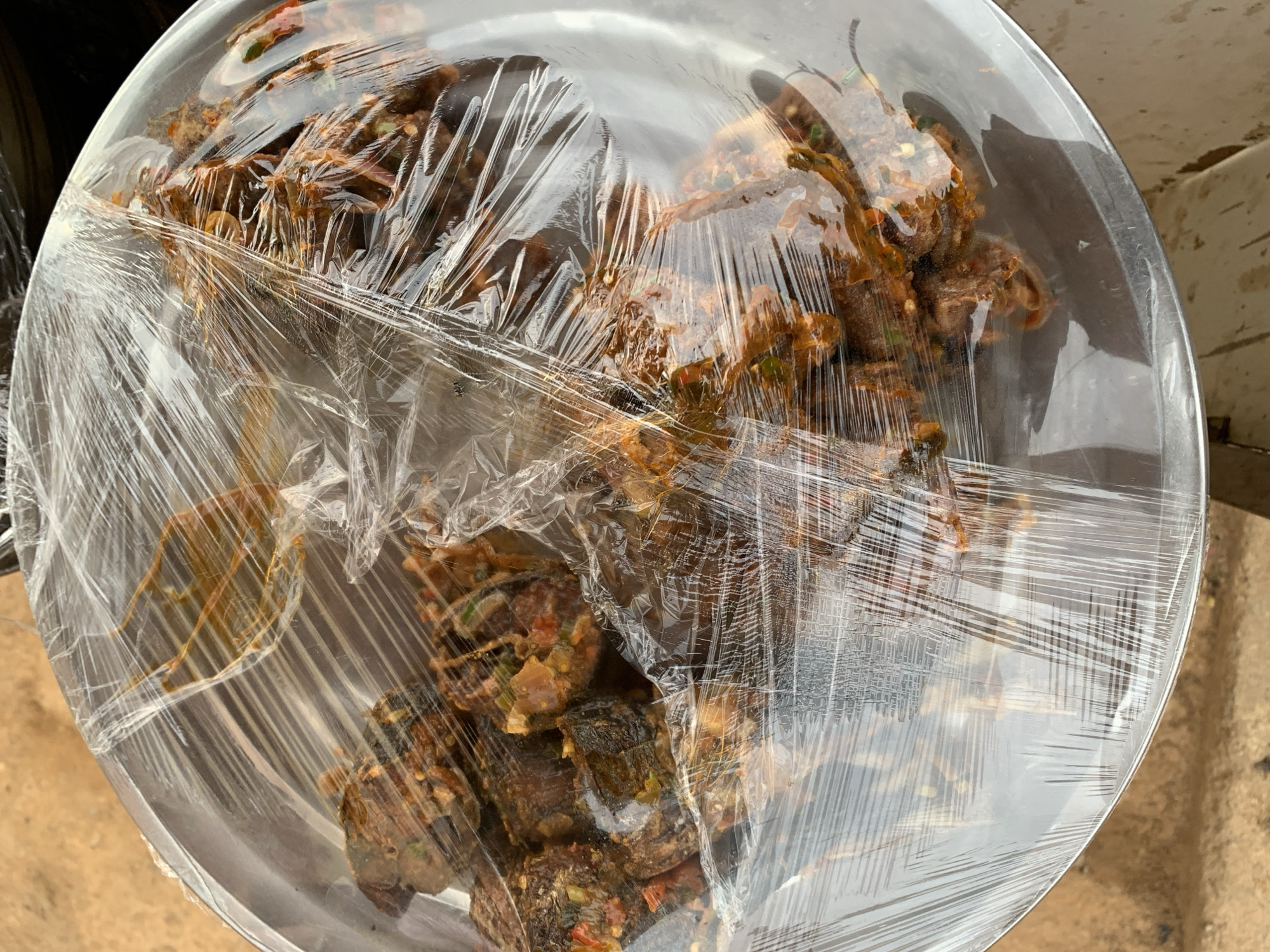 A high-angle, close-up shot shows a large metal plate filled with dark-colored food, covered tightly with transparent plastic wrap. The food appears to be in chunks, with visible spices and possibly vegetables mixed in. The background suggests an outdoor setting, possibly in Rafin Par, Nigeria. Part of a tire and an indeterminate surface are visible near the plate. The image lacks people.