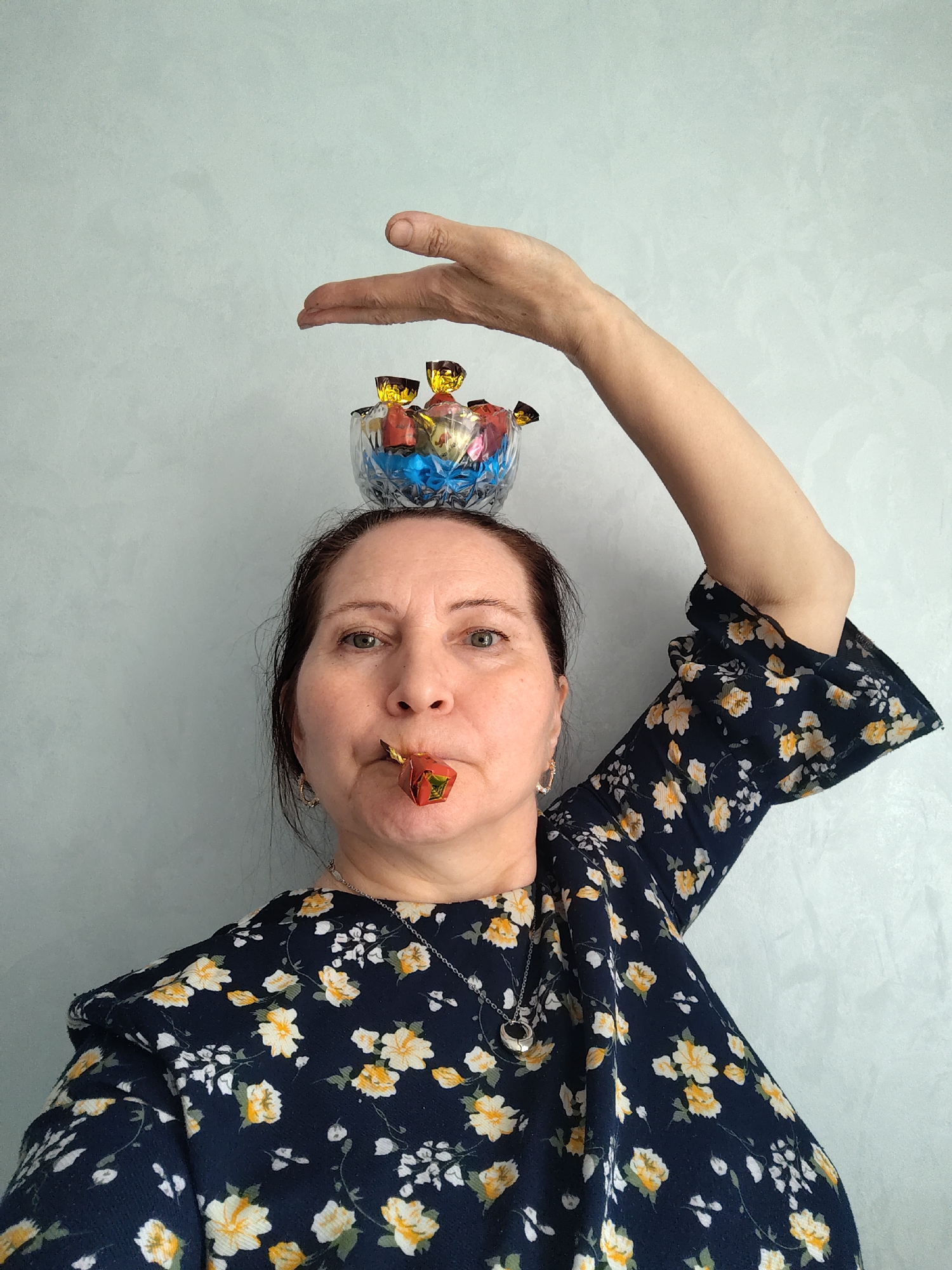 A woman stands in front of a light gray wall. She is wearing a navy blue floral dress with white and yellow flowers. She has a small clear glass bowl filled with wrapped candies balanced on her head. Her right arm is raised above the bowl with her hand open as if gesturing. She also has a wrapped candy in her mouth. She is wearing a necklace with a circular pendant and hoop earrings. The location is likely Sarapul, Russia, based on visual context.