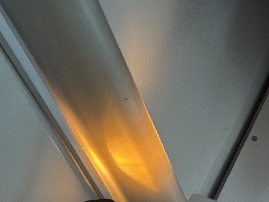 The image depicts an indoor ceiling viewed from a low angle. A rectangular, light-colored overhead light fixture, likely a fluorescent or LED strip, spans diagonally across the upper and middle portions of the frame. The fixture emits a bright, warm orange-yellow light that highlights its own surface and reflects onto the adjacent light gray ceiling. Small dark specks are visible on the illuminated section of the fixture. A dark, out-of-focus form is partially visible at the far left edge of the frame. No discernible individuals, actions, or specific environmental features are present within the image to determine a precise location in the city of Gryazi, Russia.