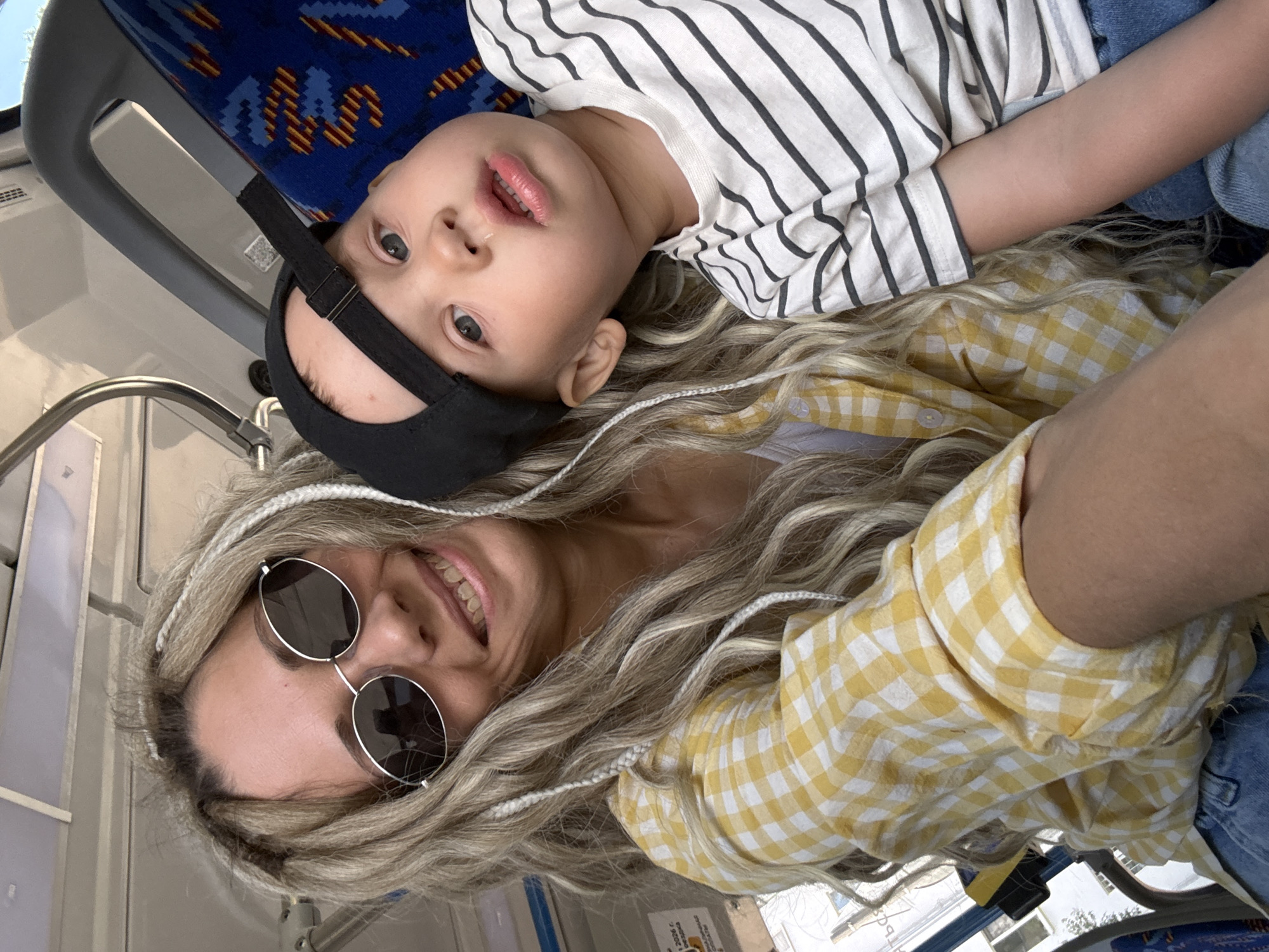 A young child wearing a black baseball cap and a striped shirt is lying next to a smiling woman wearing round sunglasses and a yellow plaid shirt. They appear to be on public transportation with blue patterned seats. A window and handrails are visible, suggesting the setting is likely Заволжье, Russia.