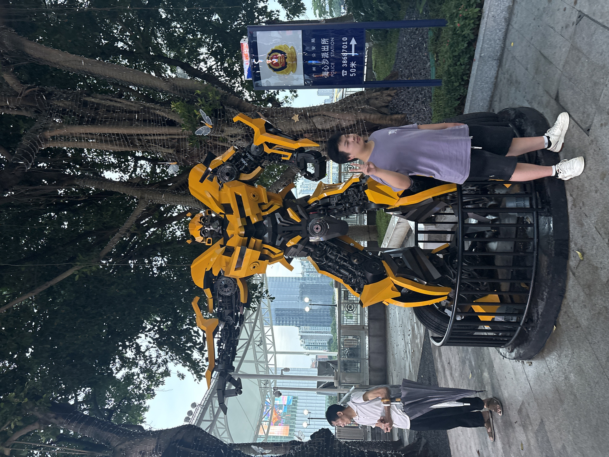 A person stands next to a large, yellow Transformer robot sculpture in an outdoor setting. The robot appears to be Bumblebee from the Transformers franchise, with intricate mechanical details and its signature yellow and black color scheme. The person, wearing a purple t-shirt and black shorts, is posing with a peace sign. Behind them, a signpost indicates "Police Station" in both English and Chinese, with a phone number and distance. Trees with dense foliage are visible in the background, along with a cityscape featuring tall buildings. Another person is seen further down in the frame, walking away with an umbrella. The lighting suggests it might be late afternoon or early evening, and the ground appears slightly damp, hinting at recent rain or a humid environment. The overall scene is a blend of urban environment and a popular culture icon.