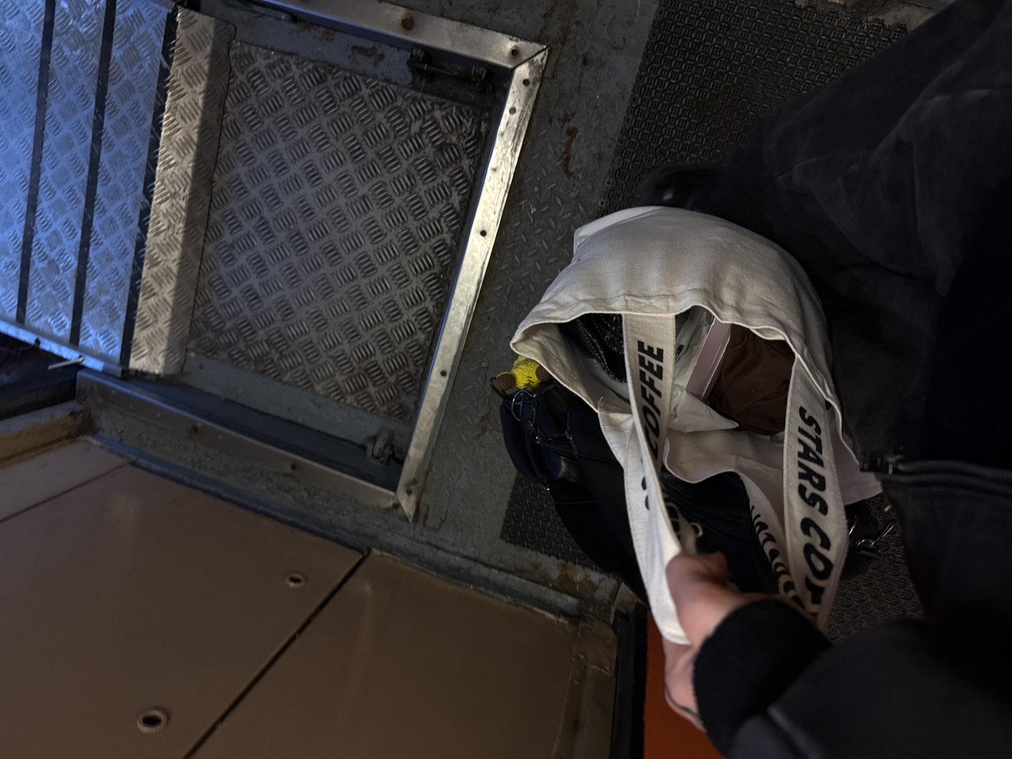 The image shows a person holding a "STARS COFFEE" tote bag, standing on a floor with different textures and levels. The floor consists of a diamond plate metal surface, a textured gray surface, and a flat, light brown surface. A metal-framed hatch is visible on the floor. The person is wearing a dark jacket and holding the tote bag with their hand. The bag contains various items. The location is suggested to be in Saint Petersburg, Russia, based on the "STARS COFFEE" tote bag, a chain that replaced Starbucks in Russia.