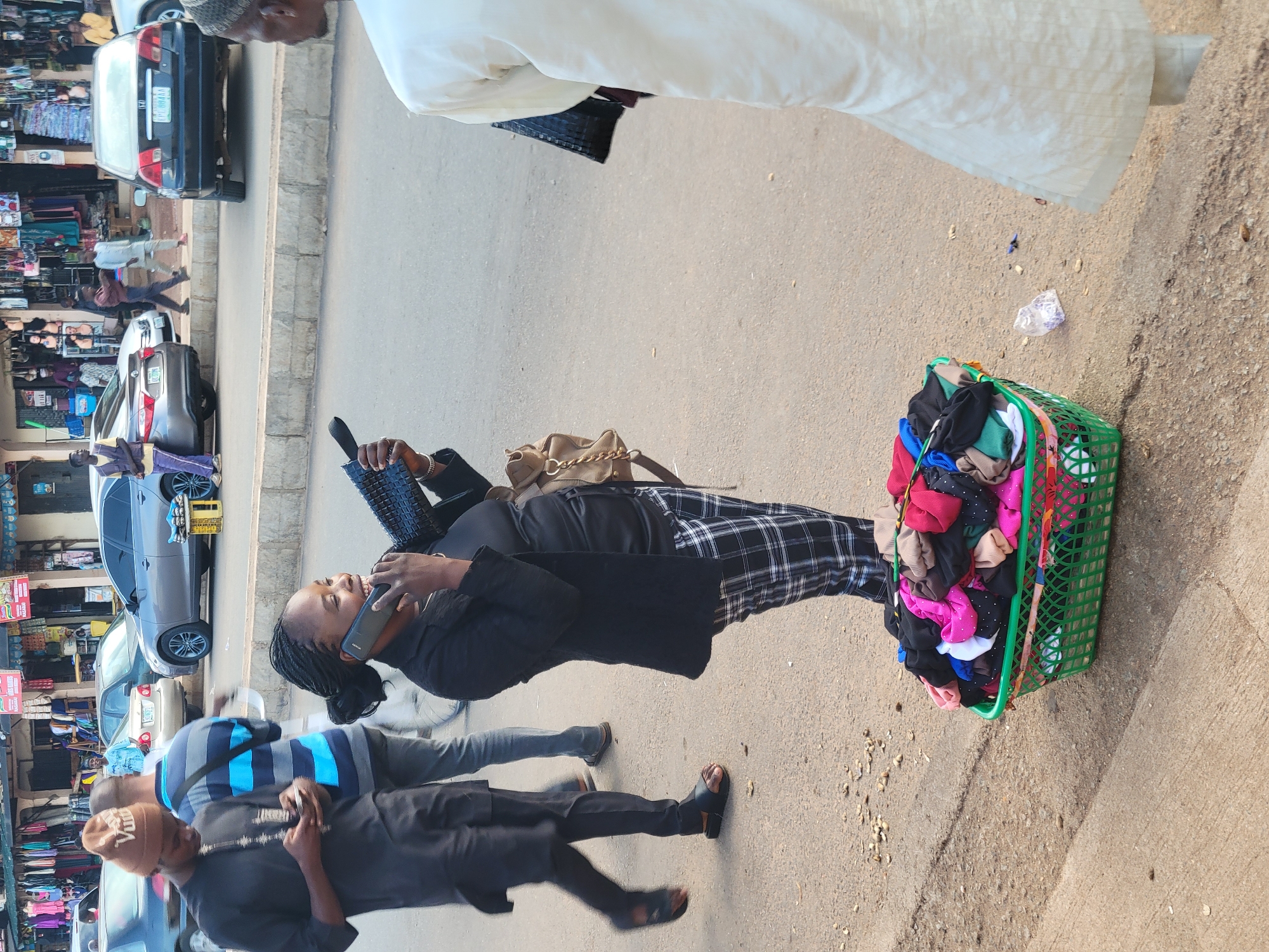 A woman with braided hair is talking on her phone while standing next to a green basket filled with colorful folded clothing. She is wearing a black top and plaid pants. A beige purse with a gold chain strap hangs on her left shoulder. The scene appears to be an outdoor market or street with cars parked along a sidewalk and shops in the background. Another person, wearing a blue and gray striped shirt, is visible walking in the foreground. The lighting suggests it is daytime. The woman's expression is focused on her phone conversation. The basket contains a variety of clothing items, including socks and possibly underwear. The ground is paved, and there are some small pebbles scattered on the surface. The overall setting suggests a busy marketplace.