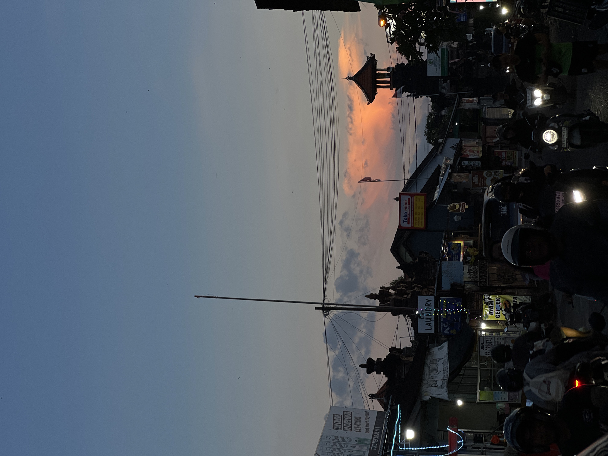 This image depicts a bustling street scene in Denpasar, Indonesia, captured during dusk or early evening. The sky transitions from a soft, warm orange and pink hue on the right, indicative of a sunset, to a darker blue and grey on the left, with scattered clouds.
The foreground and right side of the image are filled with numerous motorcycles, many with their headlights illuminated, suggesting active traffic or commuters. Several individuals, some wearing helmets, are visible on these motorcycles. Along the street, a dense array of commercial buildings and shops line the road, adorned with various signs and advertisements, some brightly lit. Overhead, a complex network of power lines extends across the frame, connected to tall utility poles.
A prominent traditional Balinese temple structure, characterized by its distinctive multi-tiered roof, is silhouetted against the sunset-lit sky, serving as a cultural landmark. Streetlights and vehicle lights contribute to the evening ambiance.
Visible text on various signs includes "TOKO MEKAR JAYA 1", "CLA'S LAUNDRY", and a sign advertising "AYAM GEKREK" (a popular Indonesian fried chicken dish). Other text states "JUAL ES CRISTAL", "Toko mini" with "Rp. 5000", and promotional phrases such as "Drink Smart Play Hard" and "LOW CALORIE NO ADDED SUGAR 4.8% ALCOHOL". Partially visible license plate numbers like "6650 AEA" and "DE 51754" are also present. The scene conveys the lively, everyday atmosphere of an Indonesian urban area as day turns to night.