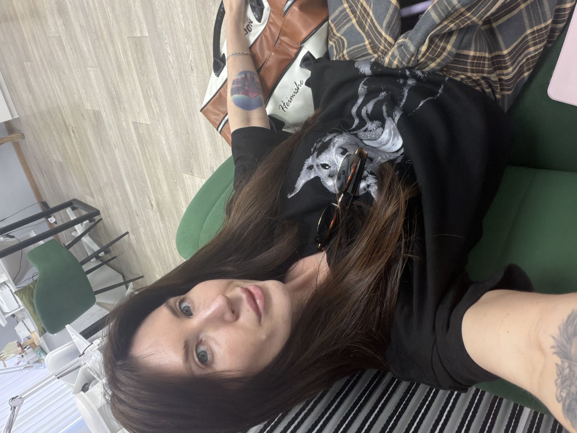 A woman with long brown hair and blue eyes is lying down, taking a selfie. She's wearing a black shirt with a wolf graphic and has sunglasses tucked into the neckline. Her left arm is visible, displaying a colorful tattoo and a silver bracelet. She is reclining against a green surface, possibly a couch or chair. A white and brown bag with "Heimische" printed on it, along with a plaid shirt, are also on the surface. In the background, there's a glimpse of a room with office furniture. The presence of the Heimische bag might indicate a location in Perm, Russia, as that is a brand often associated with the city.