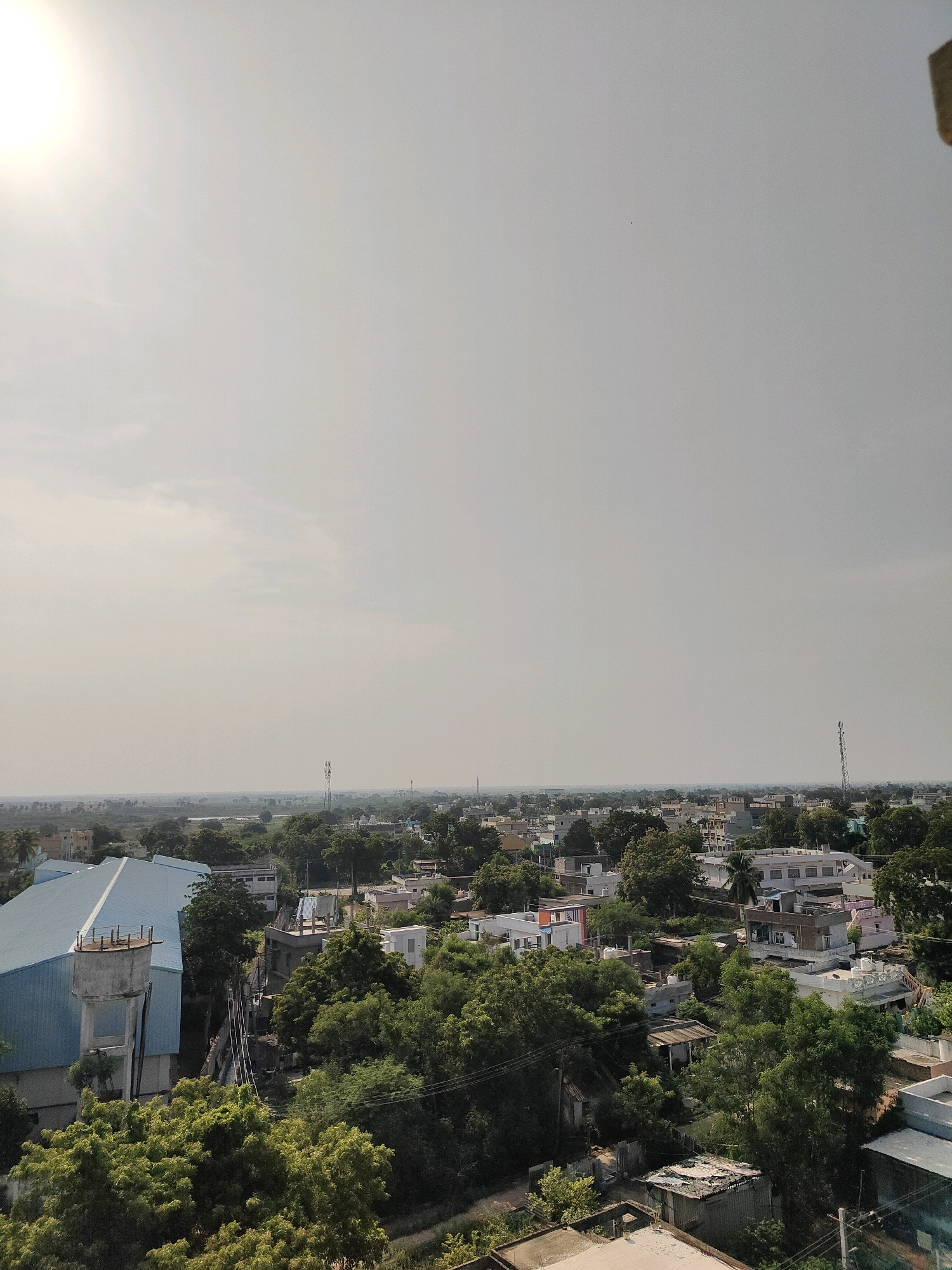 This image captures a panoramic view of the city of Medarametla, India, under a hazy sky. The sun is prominently visible in the upper left corner, its light diffused by atmospheric conditions. The cityscape features a mix of residential buildings and a large, light blue-roofed industrial building on the left, which also has a water tank attached to its side. Numerous trees are scattered throughout the scene, interspersed with houses of varying sizes and colors, including white, pink, and beige structures. Several telecommunication towers are visible in the distance, indicating infrastructure development. The overall impression is that of a moderately developed urban or semi-urban area with a warm, overcast sky.