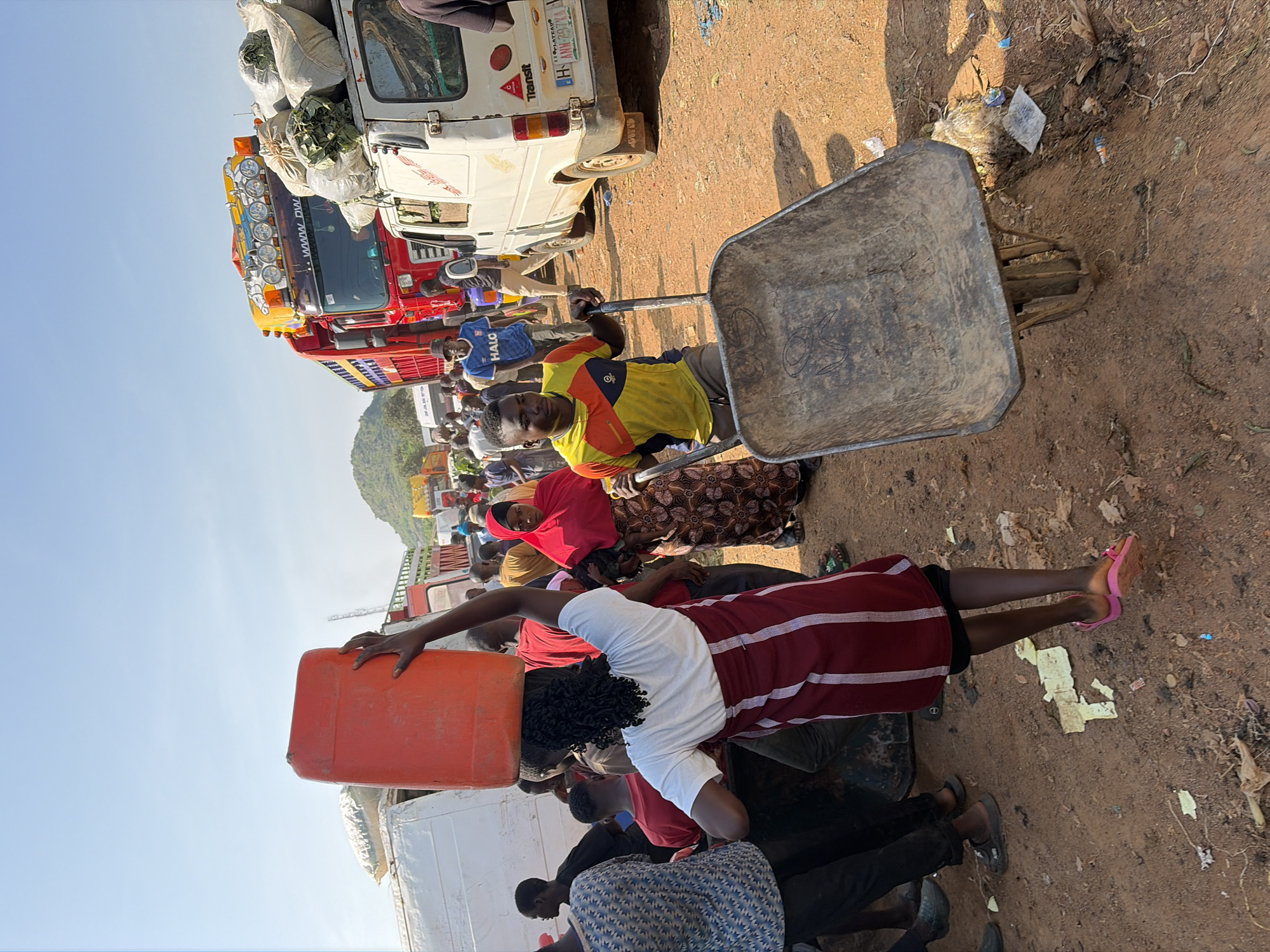 In Gura Top, Nigeria, a bustling marketplace is depicted under a bright sky. A woman, her back to the camera, carries a large orange plastic container on her head, her dark curly hair visible beneath it. She is dressed in a white t-shirt and a maroon skirt with white vertical stripes. Beside her, a man in a yellow and orange t-shirt and brown trousers pushes a weathered metal wheelbarrow. In the background, several large trucks, including a red and yellow one with numerous lights on its roof, are parked, some laden with large bags of produce. White vans are also present. The ground is covered in reddish-brown dirt, littered with debris. A crowd of people, dressed in various clothing, mills about the scene, suggesting active commerce. The environment appears dusty and somewhat unorganized, typical of a busy outdoor market.