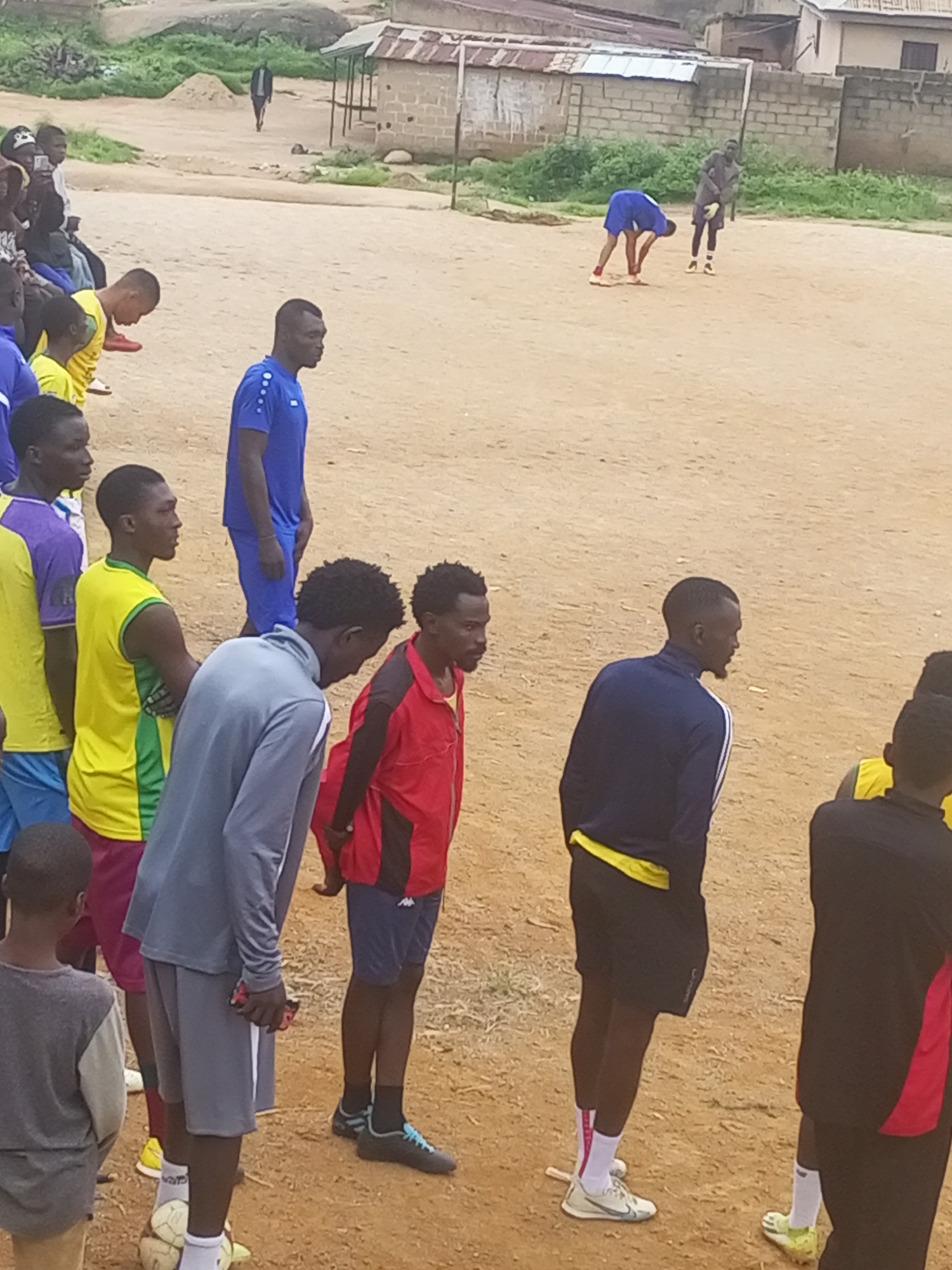 A group of young men are gathered on a dirt field, appearing to be engaged in or preparing for a football (soccer) training session. In the background, there are low-rise buildings with corrugated iron roofs, characteristic of some residential areas in Jos, Nigeria. The ground is a reddish-brown earth, with some sparse green vegetation visible in patches.
Several individuals are dressed in athletic wear. One person in a bright blue shirt and shorts is bending down, possibly tying their shoelaces. Another individual in a grey tracksuit is standing with their hands clasped, looking downwards. Others are in various football jerseys and shorts, some in yellow and green, others in blue. There's a sense of casual gathering and activity, with some individuals looking at each other or towards the field. The lighting suggests it is daytime, likely overcast or late afternoon, given the soft shadows. The overall scene depicts a common urban sports activity in a developing area.
