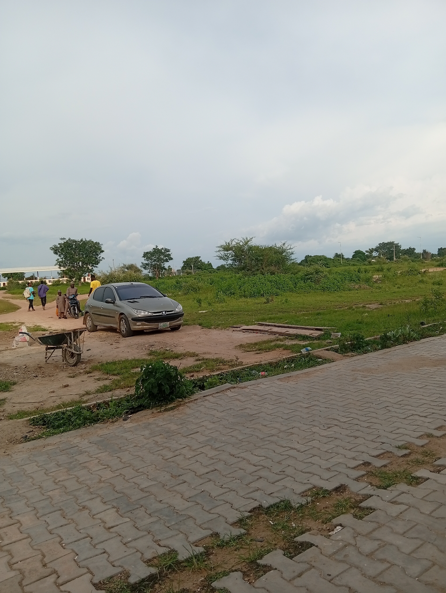 In Yali, Nigeria, on a cloudy day, a gray Peugeot 206 is parked on a dirt road. In the background, a few people are walking, and a man is on a motorcycle. A wheelbarrow is positioned in the foreground to the left, with a bag of material inside. The area is characterized by dry ground, green grass, and scattered trees, with a paved walkway visible in the lower right portion of the image.