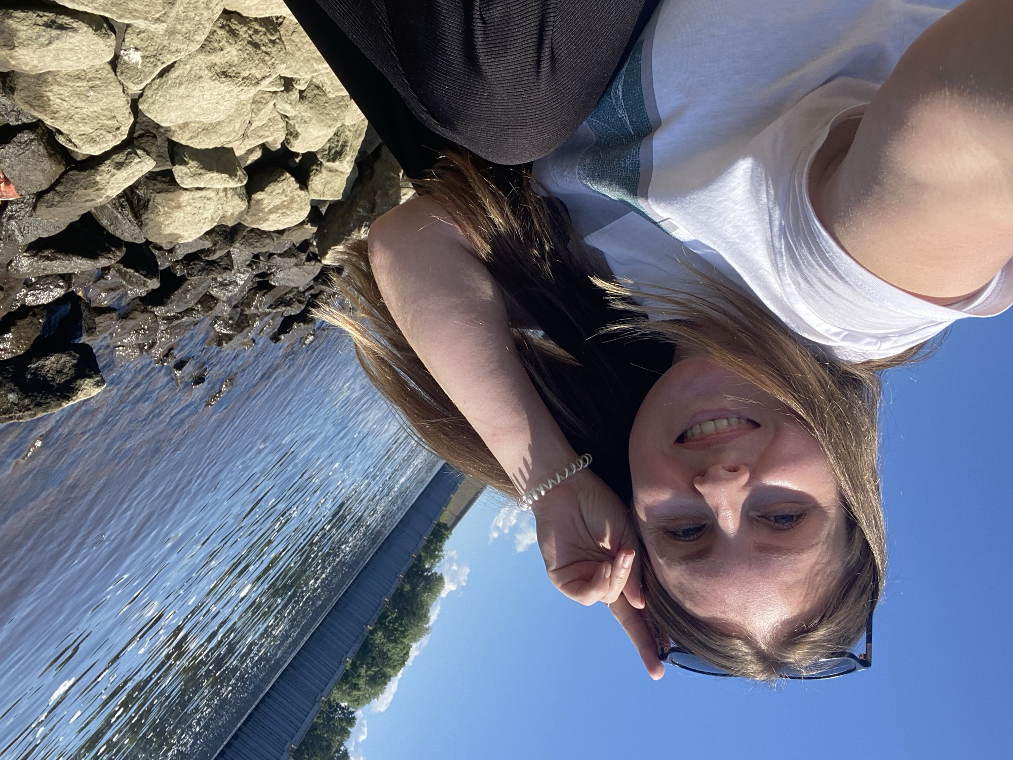 A young Caucasian woman with long, light brown hair and blue eyes is captured in a selfie. She is wearing a white t-shirt with a green and gray stripe design and black pants. Sunglasses are perched on top of her head. She is leaning on her hand, which has a pearl bracelet on her wrist, and smiling at the camera. The background features a bright blue sky, a body of water with ripples, and a dark, ribbed barrier wall. Along the edge of the water are numerous rocks, some light-colored and smooth, others dark and rough. The scene appears to be a waterfront area in Chaikovsky, Russia, possibly near the Kama River dam, given the presence of the water barrier.