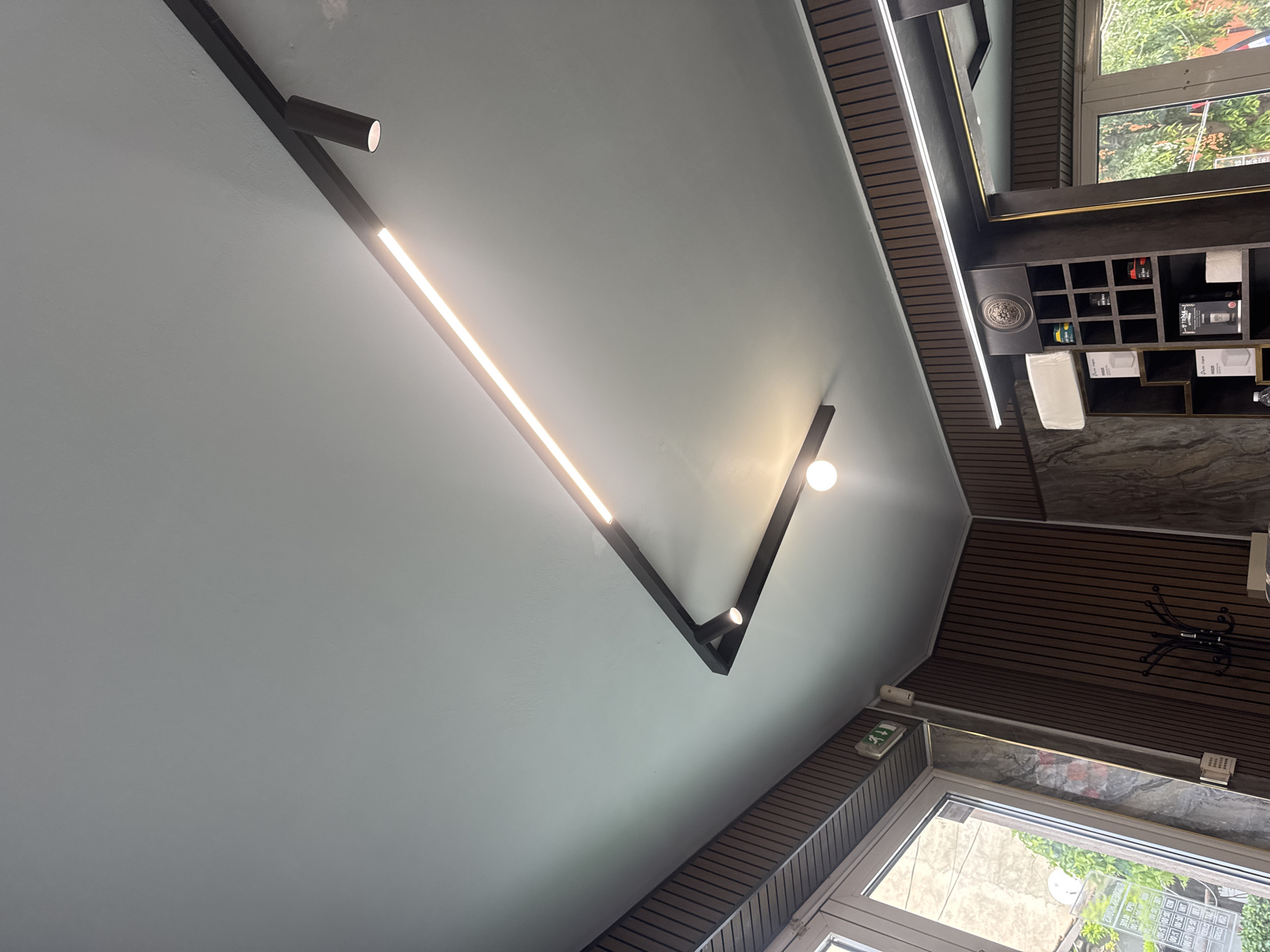 The image captures an interior scene, likely a commercial establishment such as a salon, barbershop, or store, characterized by modern design elements. No people are visible.
The dominant feature is the ceiling, painted a light grey, equipped with a striking angular black track lighting system. This system incorporates linear LED strips, emitting bright white light, and several cylindrical spotlights. A single spherical light bulb also illuminates a corner of the room.
The upper walls are clad with dark brown horizontal slatted panels, contrasting with the lighter ceiling. On the right side of the frame, a dark shelving unit with multiple square compartments is visible, holding various commercial products like bottles, boxes, and neatly folded white towels. An ornate, circular metallic medallion is affixed to the top part of this shelving.
A large window dominates the lower right portion of the image, featuring white or light-colored frames. Through the window, green foliage and what appears to be an outdoor paved area are visible, indicating it is daytime. An emergency exit sign, green with white lettering, is mounted near the window. Additional white electrical fixtures are also present on the wall. A dark, possibly decorative, metal object with curved arms hangs on the slatted wall.
Visible text includes the word "EXIT" on the emergency sign. On the window, partially obscured text in what appears to be Bulgarian can be seen, including "ПОРЪЧКИ" (Orders) and times like "08:00 - 18:00" and "09:00 - 18:00", likely indicating operating hours or service availability. The overall aesthetic is clean, functional, and well-lit.