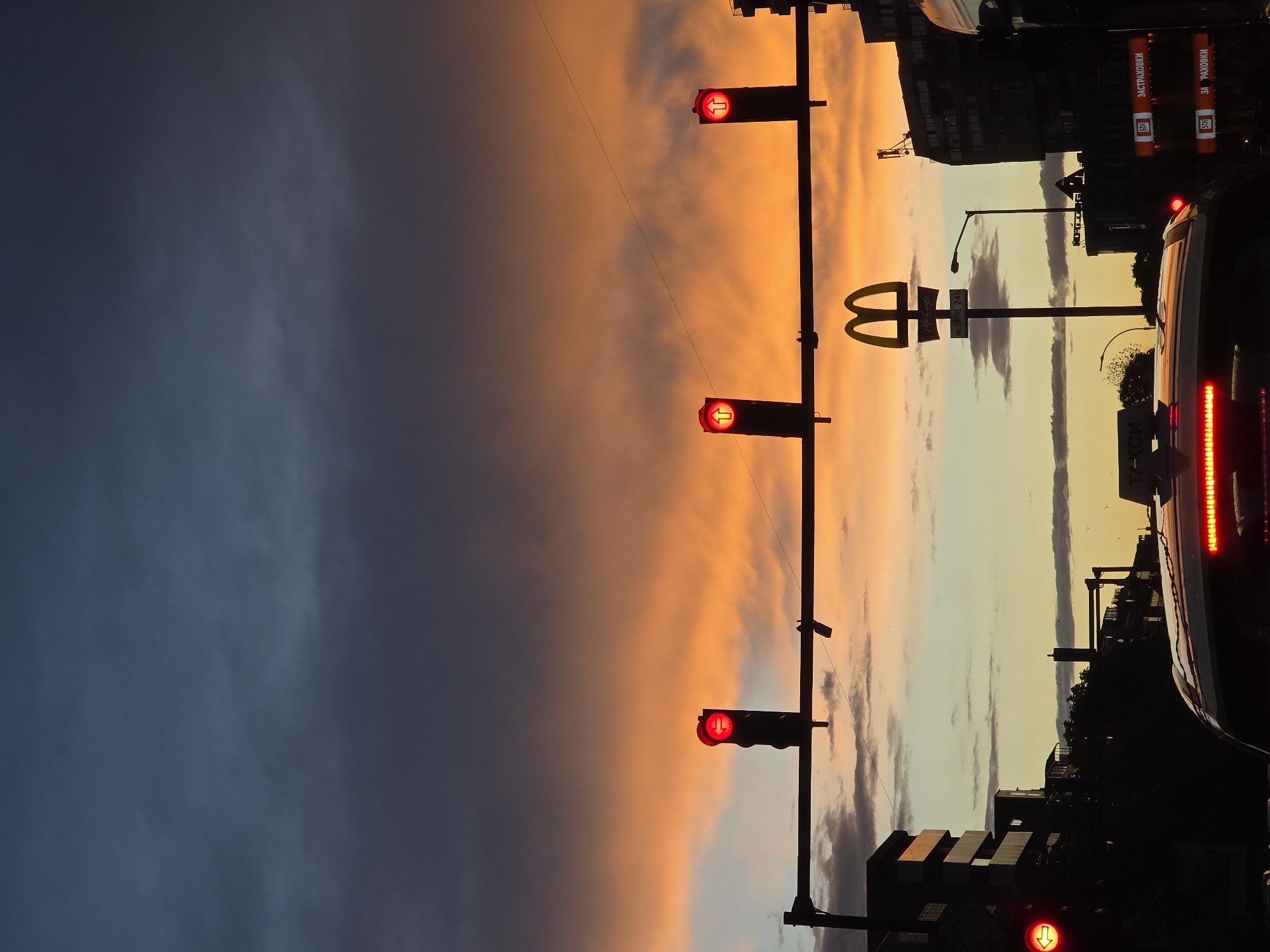 The image shows a dusk or dawn scene in what appears to be an urban environment. The sky is divided vertically into two distinct sections: the left side is a dark blue with cloud cover, while the right side transitions from orange at the top to a lighter, yellowish hue near the horizon.
A traffic light pole is centrally located in the frame, with three red traffic lights illuminated and facing the viewer. The pole also holds a McDonald's sign with the golden arches visible. Buildings and structures are visible in silhouette along the horizon line, including what appears to be an advertising billboard with Cyrillic text on it. A car's tail lights are also visible on the right side of the image.
Based on the Cyrillic text on the billboard, the location is most likely Varna, Bulgaria.