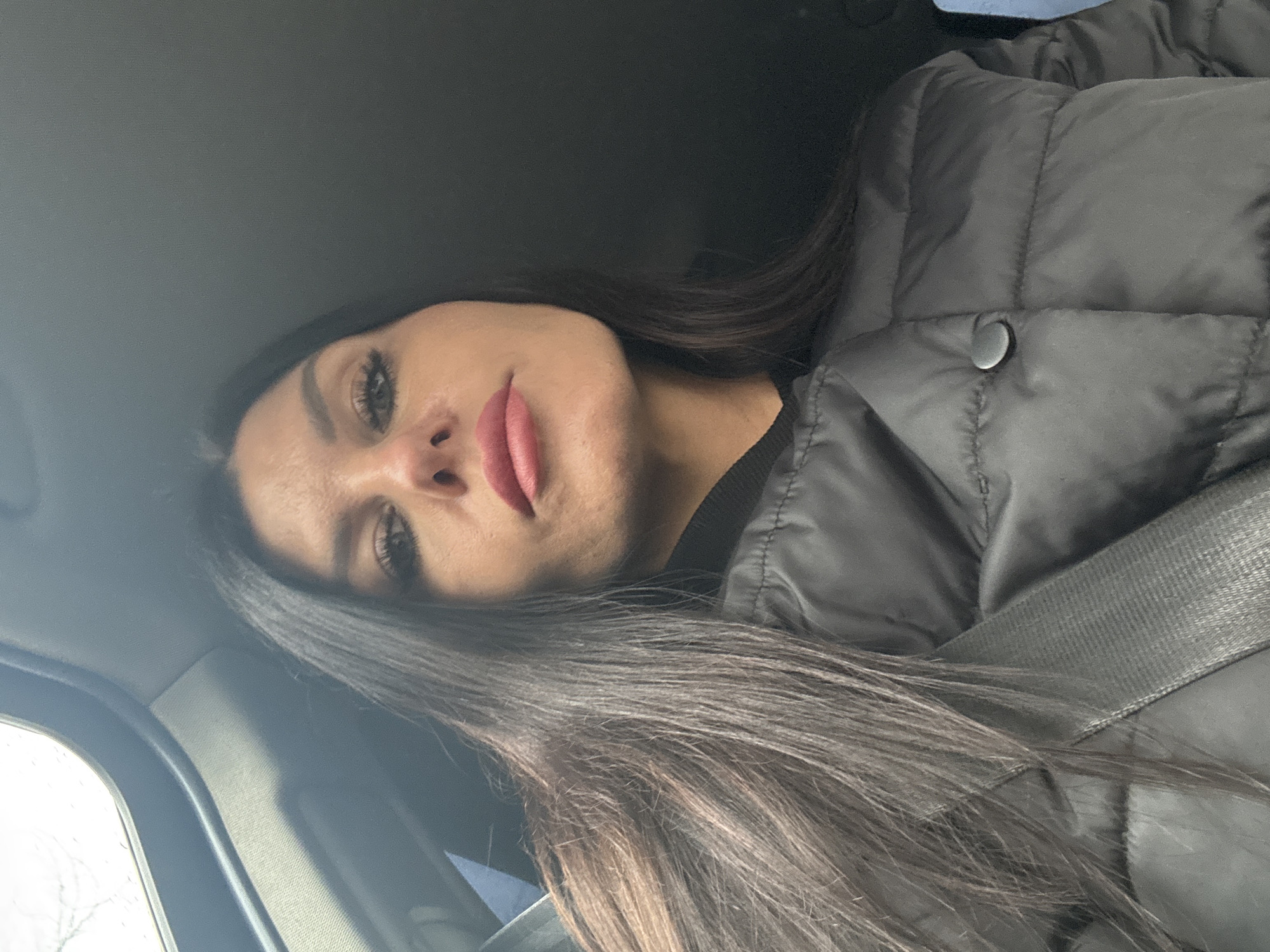 A woman with dark hair and makeup is in the passenger seat of a vehicle. She is wearing a black puffer jacket and a seatbelt. The vehicle is likely in motion. The location is unknown.