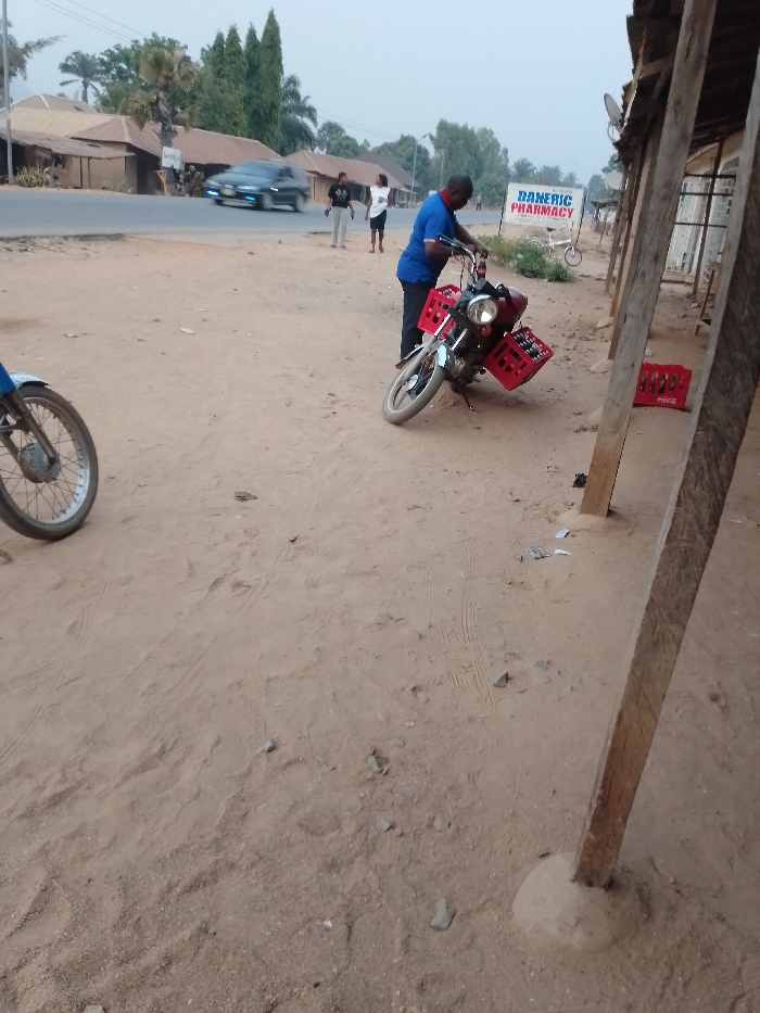 A man in a blue shirt stands next to a motorcycle laden with red crates of Coca-Cola. The bike is parked on a dirt road near a building with wooden supports. Two more people are walking on a road. In the background, there is a sign for a pharmacy, a bicycle, houses, and a moving car. A second motorcycle is partially visible on the left side.