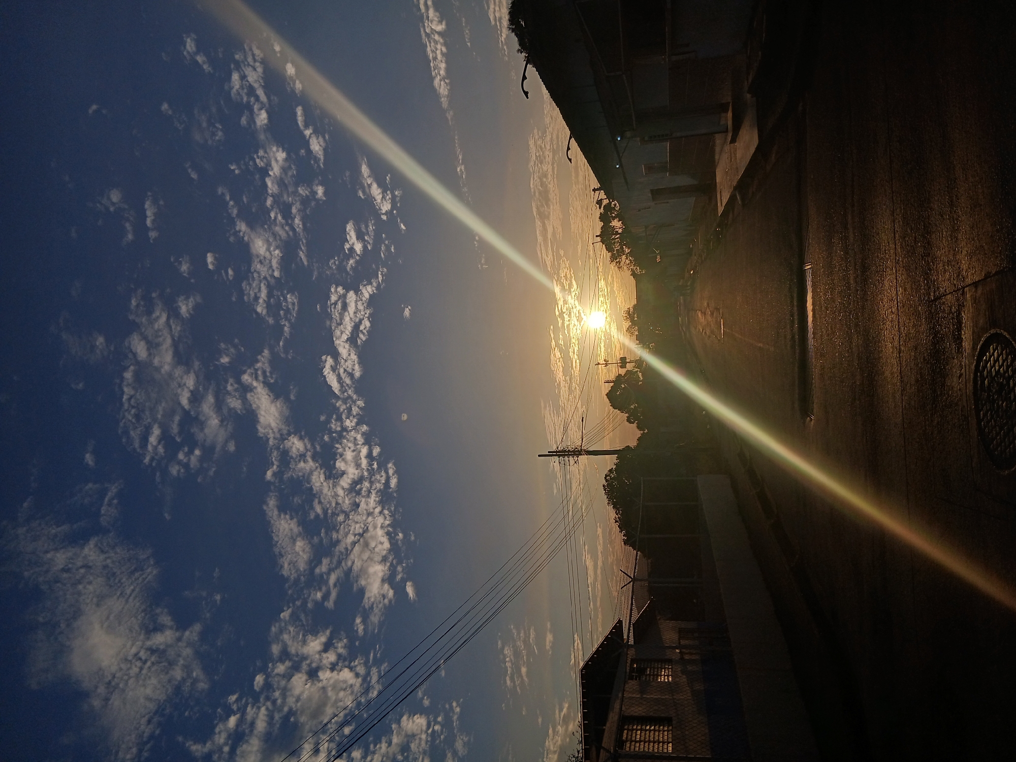 The image shows a street scene in Juan Griego, Venezuela at sunset. The sun is setting in the center of the frame, with a bright light streak emanating from it. The sky is partially cloudy, with blue visible between the white clouds. On the right side of the frame is a road, lined with buildings. On the left side of the frame are power lines and buildings. The image is taken from a low angle, looking upwards.