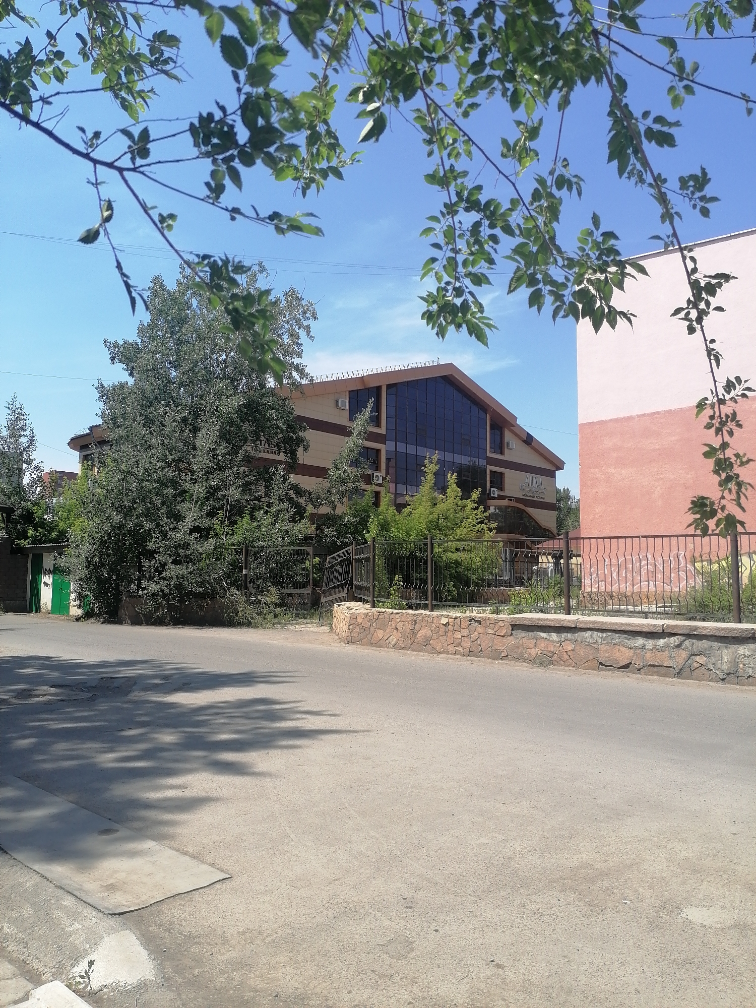 A medium shot captures a street scene on a sunny day, featuring a light pink building on the right and a beige and brown building with a large glass facade on the left. Green leaves and branches from a tree in the foreground partially obscure the view. The street is paved, with shadows from trees cast across it. A low stone wall runs along the side of the street, separating it from a grassy area in front of the buildings. A dark metal fence with decorative elements is visible in front of the beige building. Several trees and bushes are present between the two buildings and behind the fence. The sky is clear and blue.