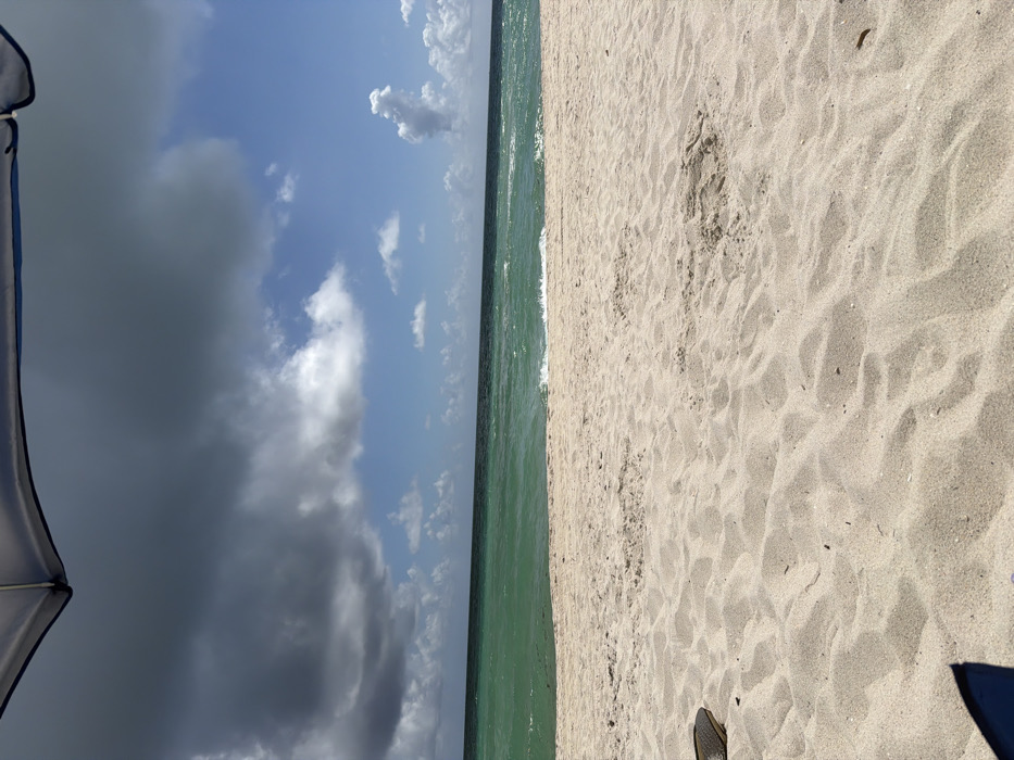 The scene depicts a beach in Miami Beach, USA, viewed from directly beneath the partial canopy of a grey and blue beach umbrella. The sky is divided, with heavy, dark grey clouds dominating the upper left quadrant and clear blue sky with scattered white cumulus clouds present towards the right. The ocean exhibits distinct horizontal bands of color, with turquoise-green water and small breaking waves near the shore, transitioning to a deeper blue-green further out towards the horizon. The foreground consists of light beige sand, displaying ripples and minor surface indentations. A dark-colored sandal or piece of footwear is partially visible on the sand in the lower left corner, and another dark, indistinct object is partially visible in the lower right. No individuals are present in the image.