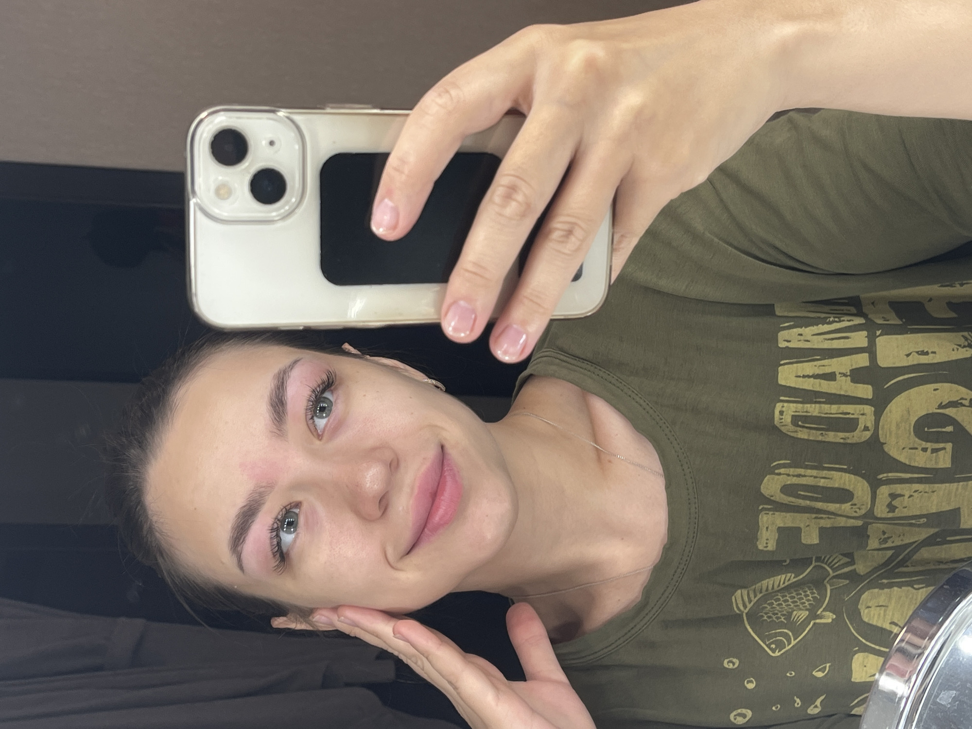 A young woman with light brown hair tied back takes a selfie in a mirror. She is wearing an olive green t-shirt with a graphic print of a fish and text. Her left hand is raised to her cheek. In her right hand, she holds a white iPhone, capturing the reflection. The background shows a dimly lit room with dark curtains. No specific location in Chaikovsky, Russia can be determined from the image.
