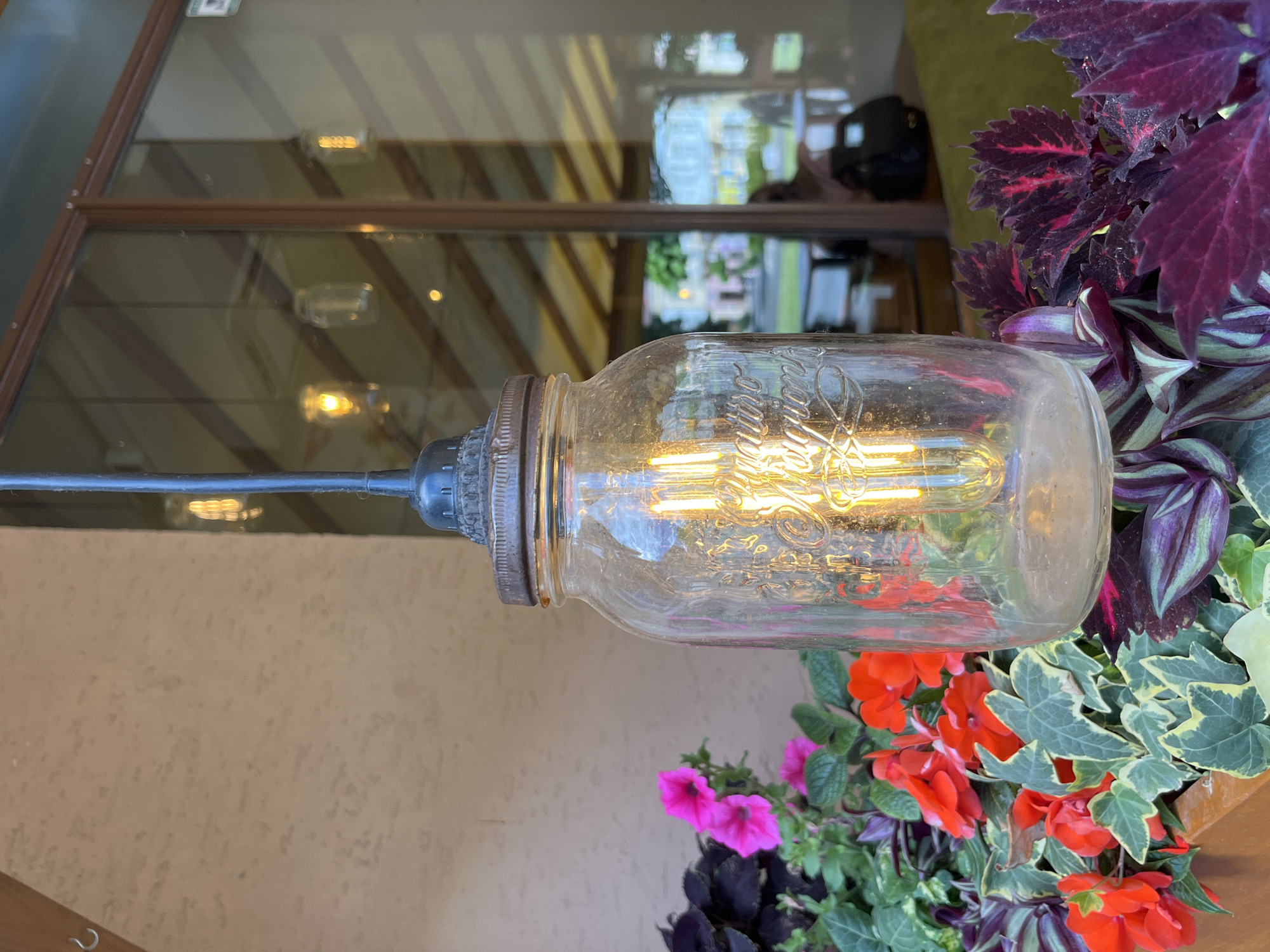 An outdoor scene features a mason jar pendant light with an Edison bulb hanging in front of a building with a textured beige wall and large windows. The light is positioned above a planter filled with vibrant red and pink flowers, along with dark purple and green foliage. Reflections in the windows show other similar lights and interior elements, including people seated at tables. The context of Drohobych, Ukraine, is not discernible from the visual information provided.