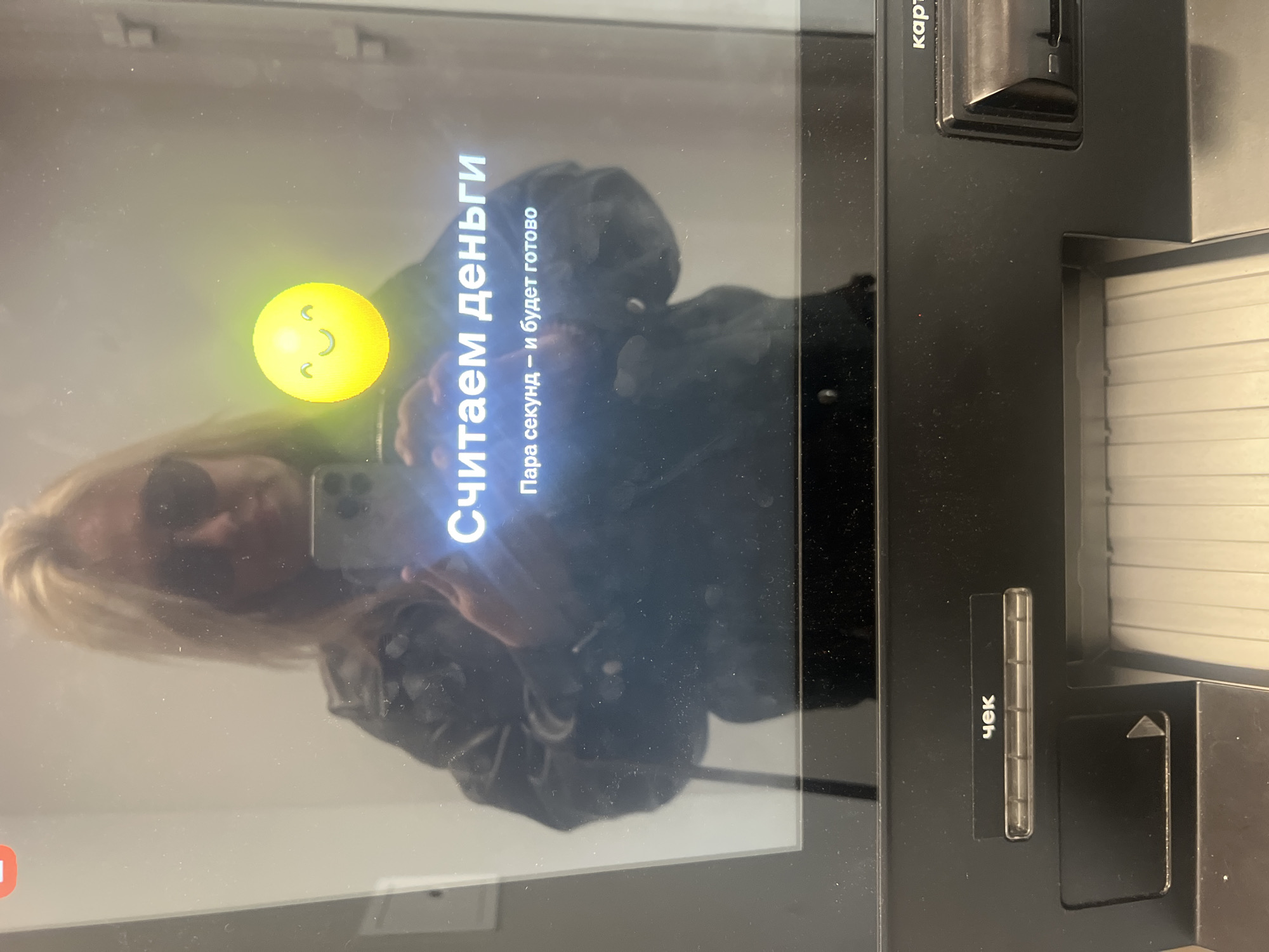 The image shows a close-up of a self-service terminal or ATM screen, featuring a reflection of a person and the machine's interface. A person with blonde hair, wearing sunglasses, and holding a smartphone (likely an iPhone) is clearly reflected on the screen.
The screen displays a bright yellow emoji of a happy, contented face with closed eyes. Below the emoji, prominent white Cyrillic text reads "Считаем деньги" (Counting money). Underneath this main message, smaller text states "Пара секунд — и будет готово" (A couple of seconds — and it will be ready). These messages indicate that a cash transaction is currently being processed by the machine.
To the right of the screen, the dark gray or black body of the ATM/terminal is visible. It features a slot labeled "карта" (card) at the top right, intended for card insertion, and another slot labeled "чек" (receipt) at the bottom right, where receipts are dispensed. The overall setting appears to be indoors, likely a bank or a commercial space in Chaikovsky, Russia, given the Cyrillic text. The reflections on the screen also reveal some dust particles.