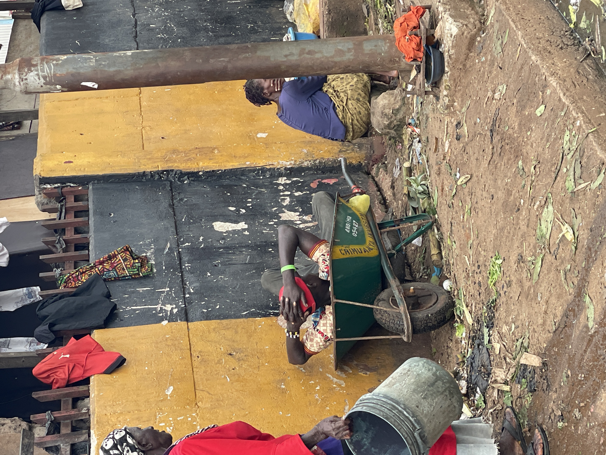 The image depicts a street scene in Onitsha, Nigeria, characterized by a weathered yellow and black wall that forms a backdrop. In the foreground, a person wearing a red garment and a headscarf stands to the left, holding a large gray bucket. A man is seen lying down in a green wheelbarrow, with his hands clasped behind his head, wearing a red cap and a patterned shirt. To the right, a woman in a purple top and a patterned skirt sits on a rock. Various items are scattered on the ground, including debris, leaves, and what appears to be trash. Clothing items are visible hanging on a wooden fence above the wall. The overall environment appears unkempt, with evidence of an urban, possibly impoverished setting.