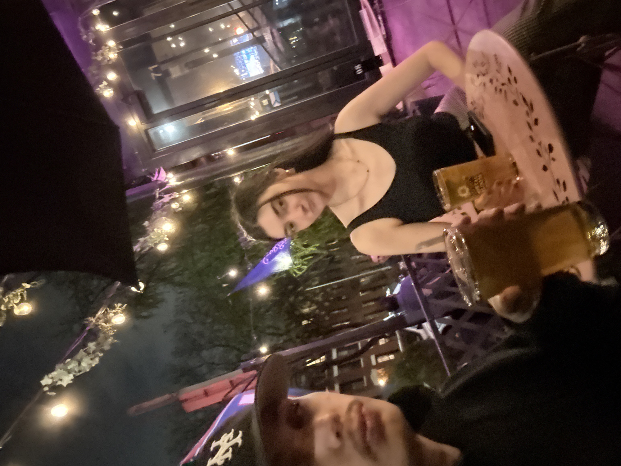 The image captures a nighttime scene at an outdoor patio in New York City, United States of America. A woman with dark hair, wearing a black top, sits at a table, holding a glass of beer. A person wearing a baseball cap is partially visible in the foreground, also holding a beer. The outdoor patio is illuminated by string lights and decorative flags, with a view of city buildings visible through the windows in the background. There are tables and chairs arranged around the patio, suggesting a restaurant or bar setting.