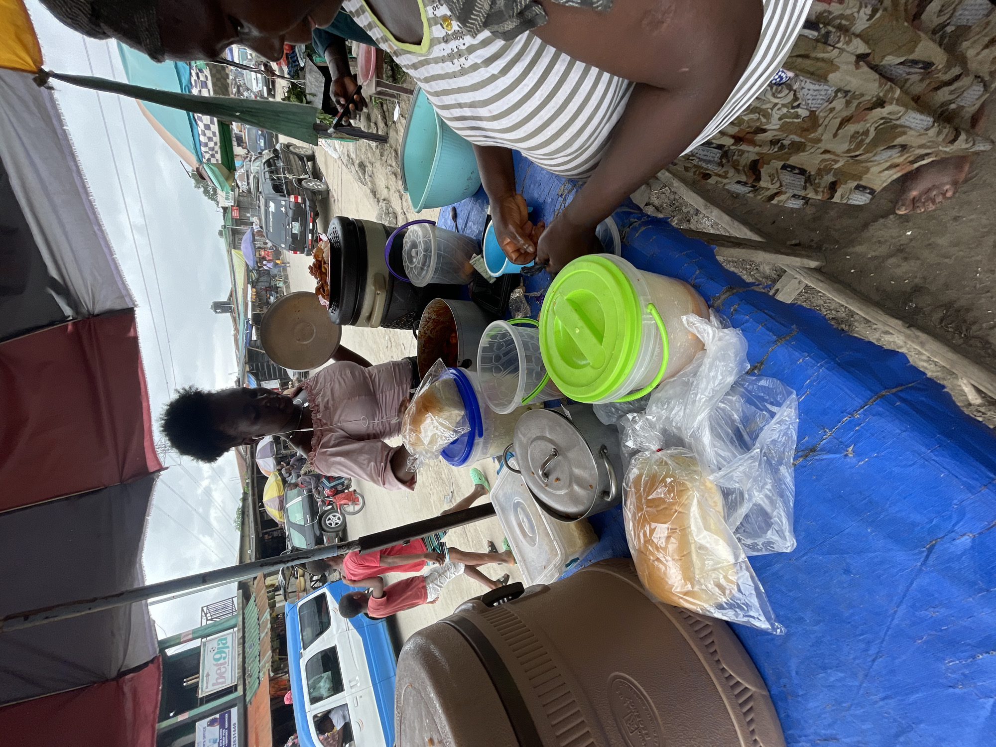 In Ado, Nigeria, a street vendor is seen under a multi-colored umbrella, displaying food items on a blue tarp-covered table. The vendor, a woman wearing a striped shirt and patterned skirt, is preparing food and interacting with customers. Several containers of food, including what appears to be stew, are visible, along with bread in plastic bags. In the background, a white and blue van, a black SUV, and motorcycles are present, suggesting a bustling urban environment. The scene depicts a typical street food setup in a lively Nigerian city.