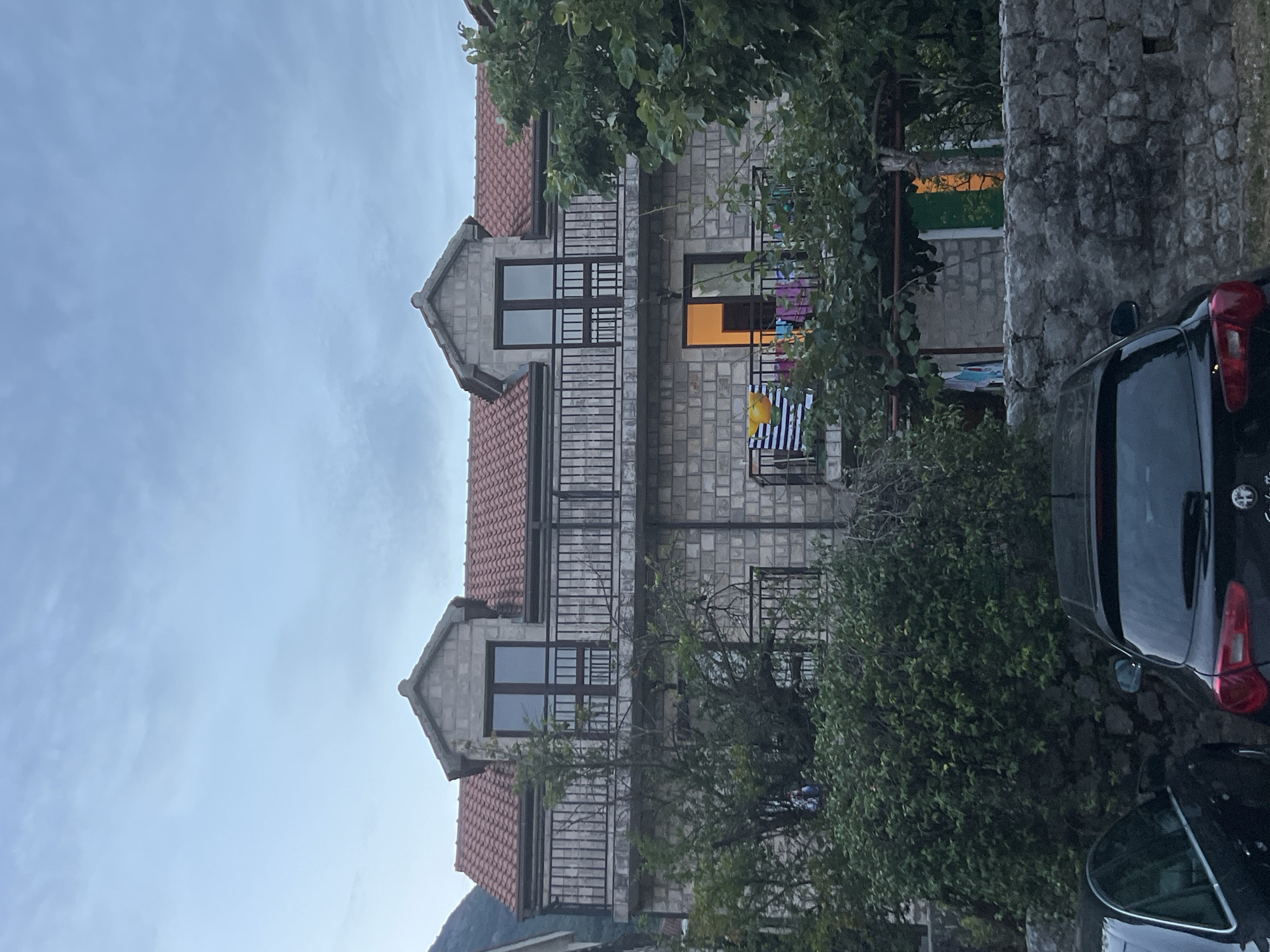 This is a photograph of a multi-story building constructed with light-colored bricks, featuring red tiled roofs. The building has multiple balconies with metal railings. On one of the balconies, laundry is hanging, including striped and colorful items. A window on the second floor is illuminated from within, casting an orange glow. Lush green foliage surrounds the building, and a stone wall is visible on the right side of the frame. A black car is parked in the foreground, partially obscuring the lower portion of the building and the vegetation. The sky appears overcast with a soft blue hue, suggesting it could be late afternoon or early evening, or simply a cloudy day. The scene is set in Baošići, Montenegro. There is no discernible text in the image.