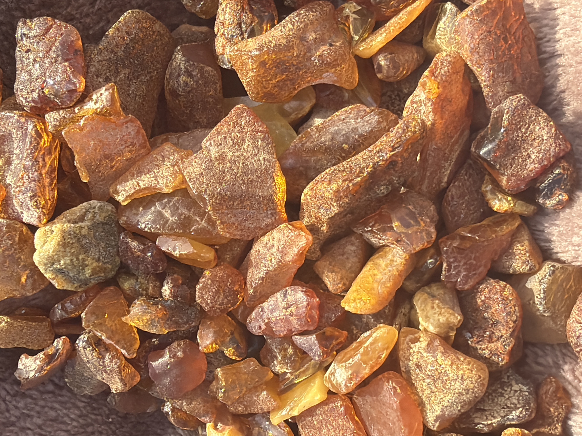 The image displays a close-up, overhead view of a collection of small, irregularly shaped amber pieces. The amber ranges in color from light yellow to deep orange and brown, with varying degrees of translucency. Some pieces appear rough and unpolished, while others have smoother, more faceted surfaces. The amber is piled on top of a textured, brown fabric. There are no people or specific landmarks visible in the image. The context of Baltiysk, Russia is not visually represented within the photograph.