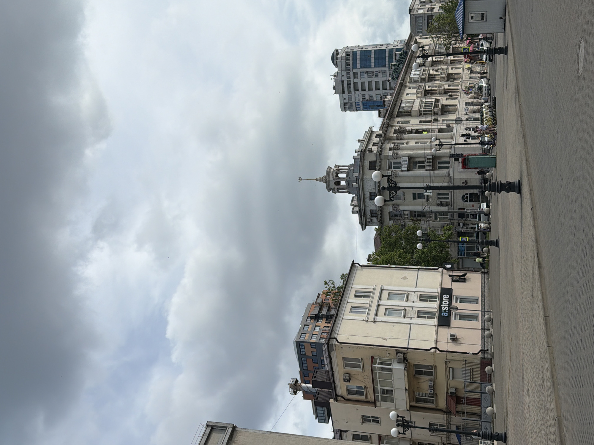 The image shows a street scene in Novorossiysk, Russia, under a cloudy sky. In the foreground, a paved pedestrian area with bollards and streetlights is visible. Several buildings line the street, varying in architectural style and height.
To the left, a light-colored, multi-story building with balconies and visible air conditioning units is present. Above it, a more modern building with dark and orange facades is seen. A sign for "a:store" is displayed on the light-colored building.
Further down the street, a prominent, ornate building with classical architectural elements and a tower topped with a spire stands out. Adjacent to this, a tall, modern skyscraper with a blue-tinted glass facade dominates the skyline.
Streetlights are present throughout the scene, and a small blue and white kiosk is visible on the right. A few people are seen walking or standing near the buildings, and several electric scooters are parked in a row. Vehicles are also present on the street, suggesting an urban environment. The overall atmosphere is overcast.