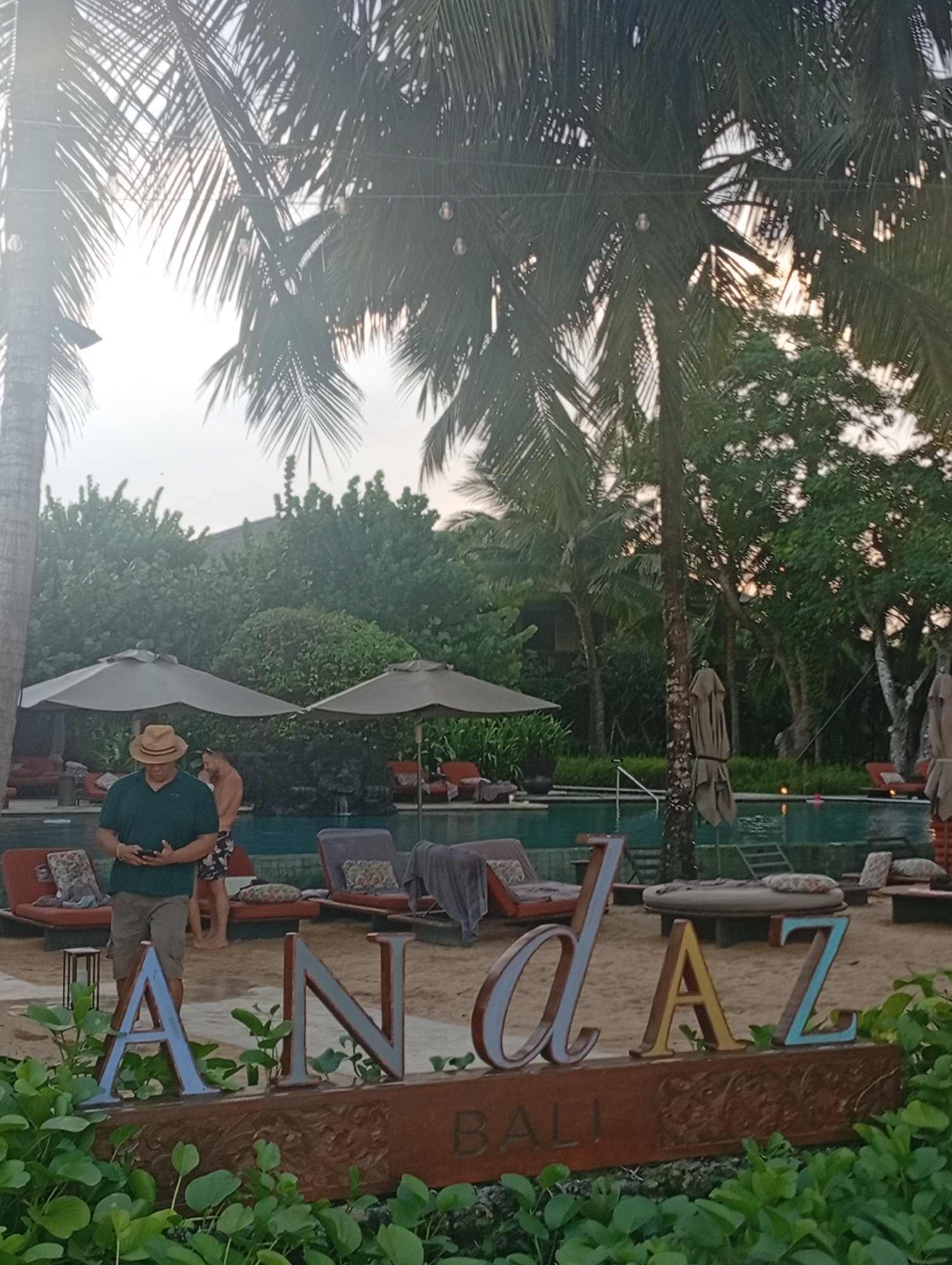 The image shows the Andaz Bali hotel in Denpasar, Indonesia. In the foreground is a decorative sign that reads "Andaz BALI." Behind the sign, there is a sandy area with lounge chairs and umbrellas. A pool of water is behind the lounge chairs. Two people are standing near the pool. Tall palm trees and other vegetation are visible throughout the scene. The lighting indicates it is either early morning or late afternoon.