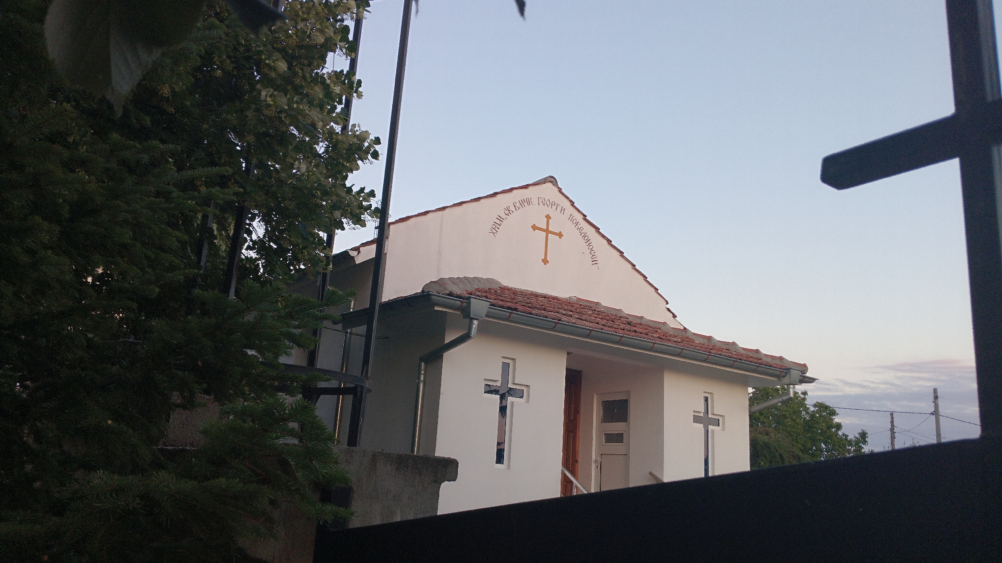 The image shows a small, white Orthodox church with a red-tiled roof, located in Dolno Izvorovo, Bulgaria. The church has a prominent orange cross on its gable, with Cyrillic inscriptions above and to the sides of the cross. There are two blue crosses visible on the facade, one above a window and another to the right of the entrance. A dark metal fence partially obscures the view of the church, and a green coniferous tree is visible in the foreground on the left. The sky is a light blue, suggesting it is daytime. The overall scene conveys a sense of modest religious architecture in a rural or semi-rural setting.