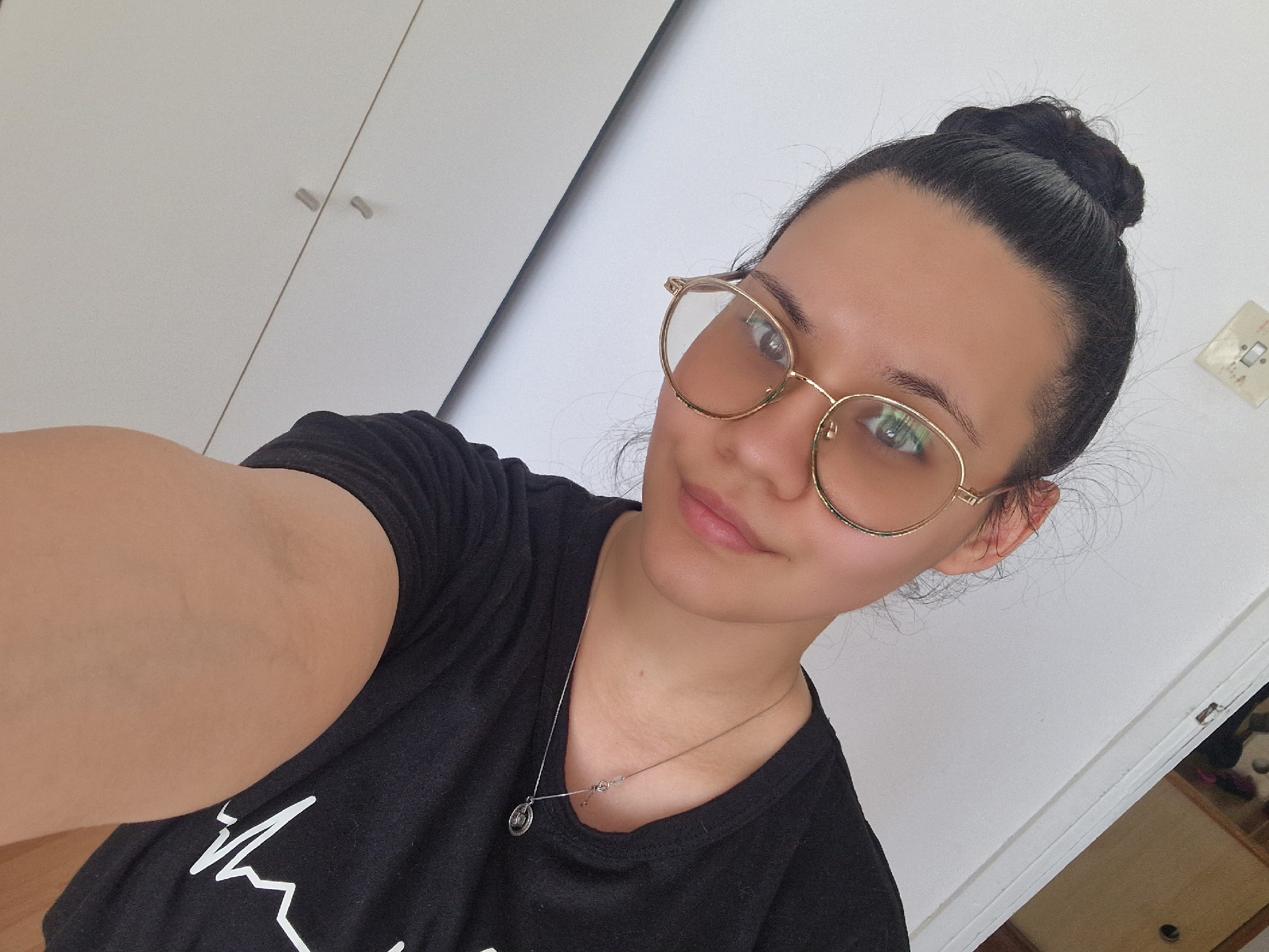 A medium shot photograph depicts a young woman indoors. She is wearing round, gold-rimmed eyeglasses, a black t-shirt with a white graphic design, and a silver necklace with a pendant. Her hair is pulled back into a bun. The background includes a white wall and white cabinets with silver handles. The presence of a European electrical outlet on the wall suggests the location may be in Europe, although there is no evidence to suggest that this image was taken in Lovech, Bulgaria. There is no indication of the specific city of Lovech, Bulgaria.