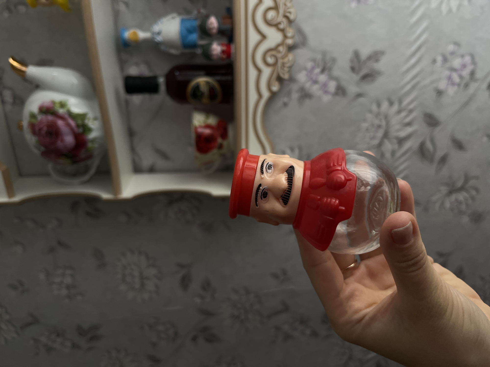 The image shows a hand holding a glass jar with a red plastic head resembling a chef, against a backdrop of a shelf containing decorative objects and floral-patterned wallpaper. The location is Мшинская, Russia.