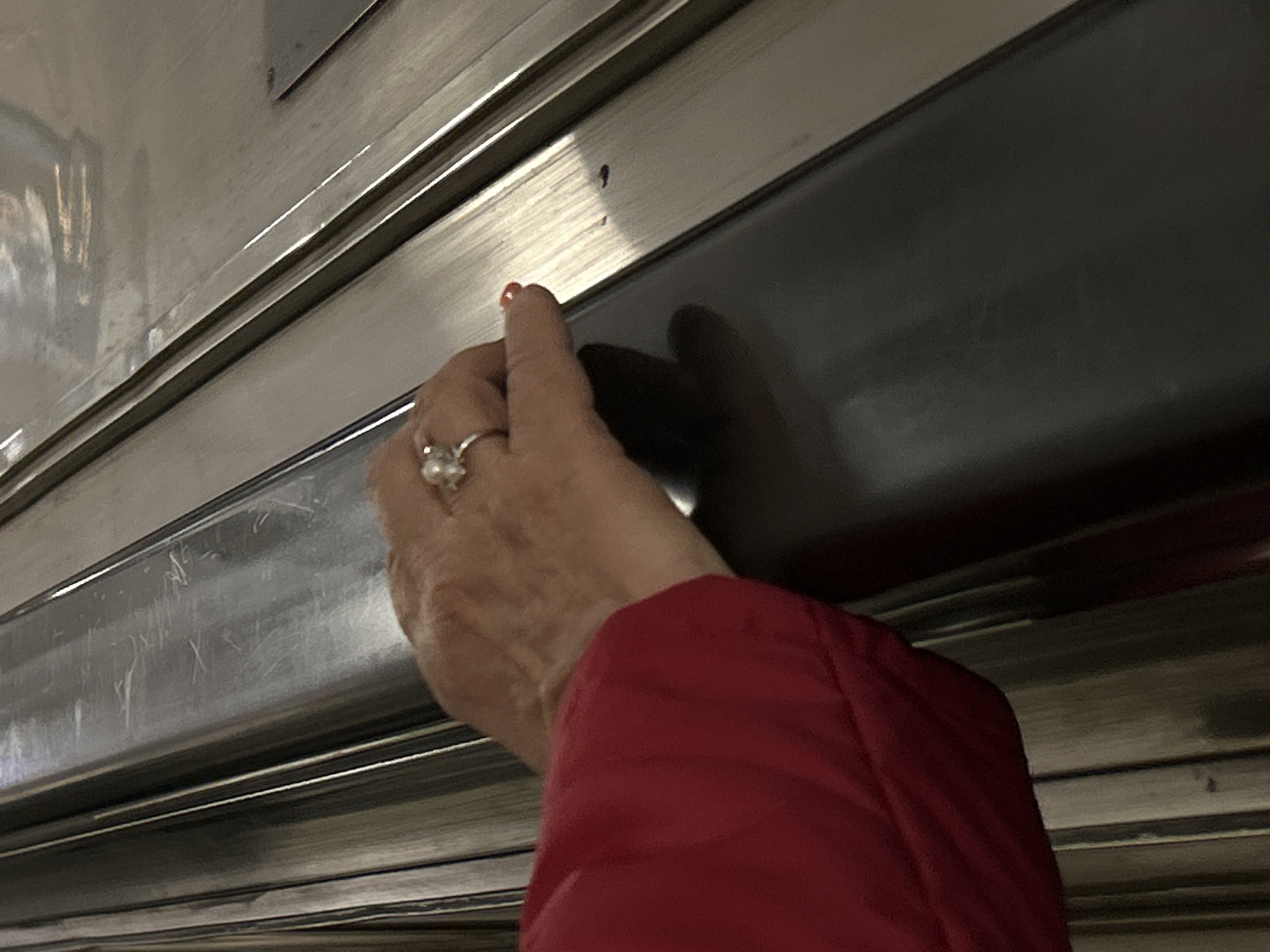The image presents a close-up view of a person's hand and lower arm within what appears to be a public transport setting. The hand, wearing a silver ring with a prominent pearl, grips a dark, horizontal handle or strip. The thumb has red nail polish. A bright red jacket sleeve covers the person's forearm, extending from the bottom right of the frame.
The background consists of metallic panels with horizontal ridges, typical of the interior design found in metro trains or other rail vehicles. These surfaces show some signs of wear and scratches. The lighting is artificial, illuminating the metallic surfaces and creating reflections. No specific time of day or external weather conditions are discernible. The activity shown is the person holding onto a support, suggesting they are standing in a moving vehicle. There is no visible text in the image. While the image itself does not provide explicit location cues, the provided context indicates this scene is set in Saint Petersburg, Russia, strongly suggesting the Saint Petersburg Metro.