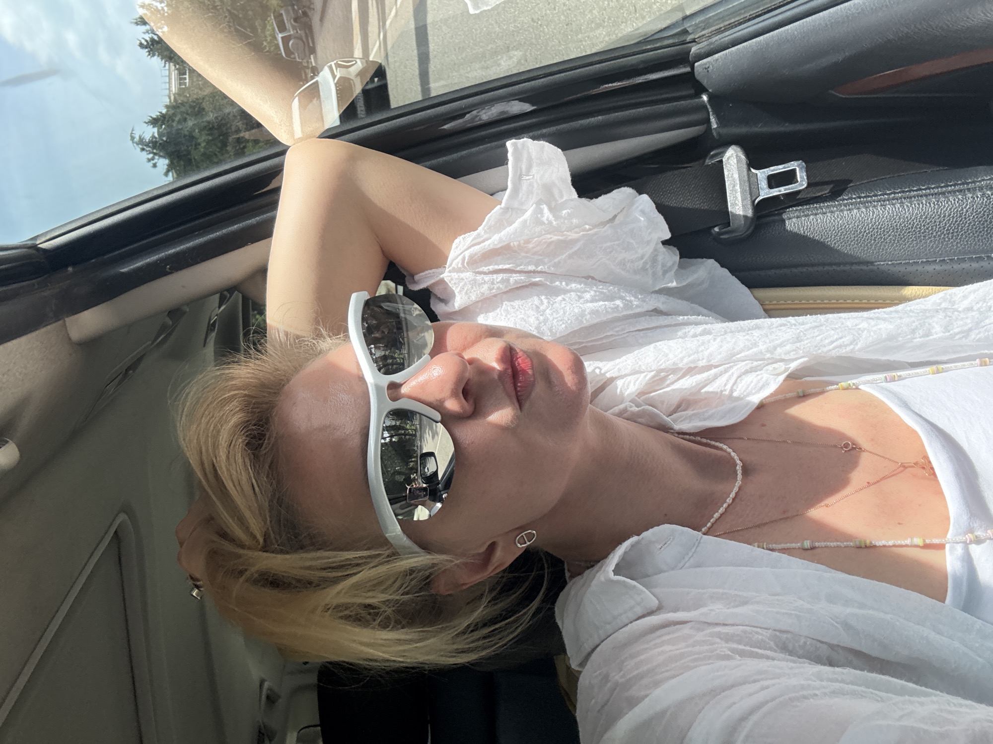 A blonde woman wearing white sunglasses and a white blouse lies in a car seat, with her head against the car door. The background shows a street scene reflected in the sunglasses. The location is Unknown, Unknown.
