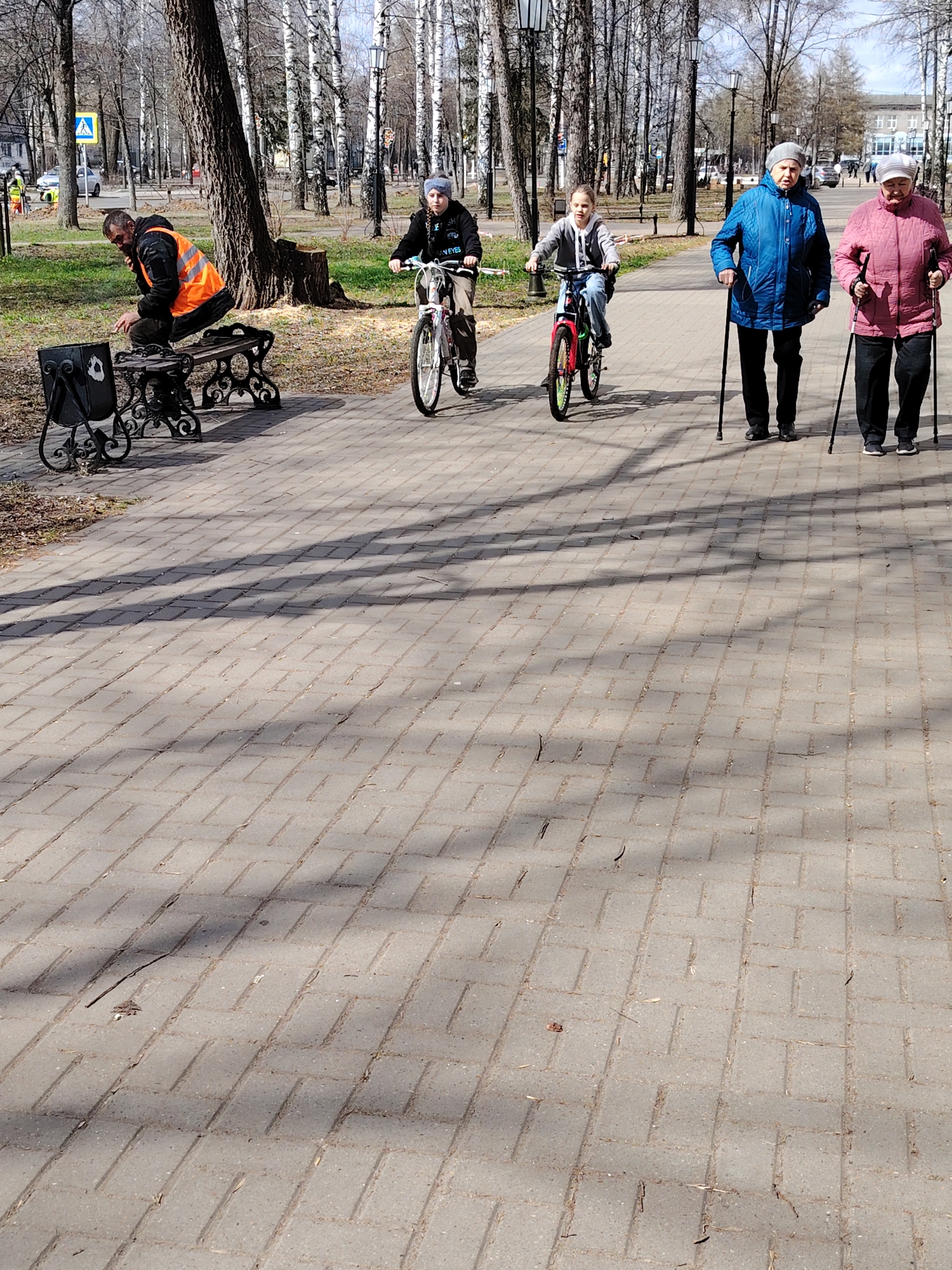 The image depicts a sunny day at an outdoor park in Chaikovsky, Russia. The ground is covered in interlocking paving stones, with tree shadows cast across it. On the left, a man wearing an orange safety vest is sitting on a bench beside a black metal trash receptacle. Two children are riding bicycles on the paved path, one with a white bicycle and the other with a red bicycle. To the right, two elderly women are walking side by side, using canes for support. In the background, there are birch trees and lamp posts, suggesting a park environment. A road sign and what appear to be workers near a vehicle can be seen in the far background.