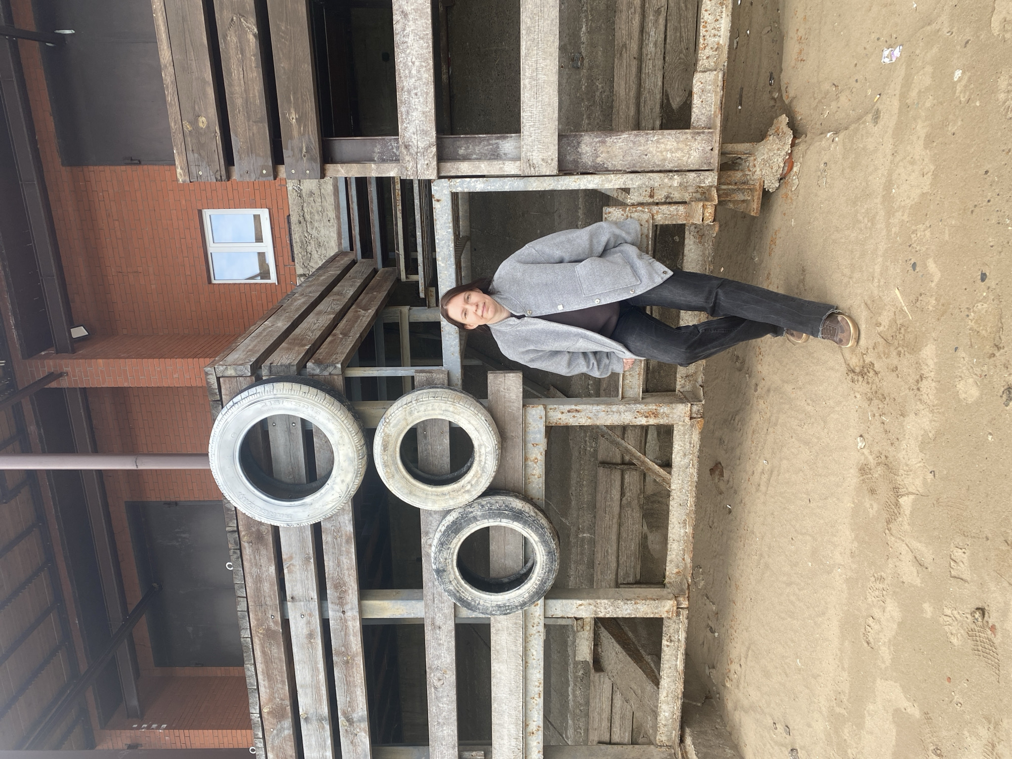 A woman stands outdoors in an industrial or utilitarian setting in Chaikovsky, Russia. She is positioned slightly to the right of the frame's center, looking directly at the camera with a neutral to slightly smiling expression. She wears a light grey button-up jacket over a dark top, dark trousers, and brown shoes.
She stands beside a large, multi-level structure constructed from weathered wooden planks and rusted metal frames, which dominates the left side of the image. Three old, dirty tires are placed on a lower platform of this structure. Text visible on one of the tires includes "215/65R16". In the background, to the left, a red brick building with a white-framed window is visible. The ground is an uneven surface of light brown dirt or sand with scattered debris. The lighting is soft and diffuse, suggesting an overcast day. The woman appears to be posing for a photograph.