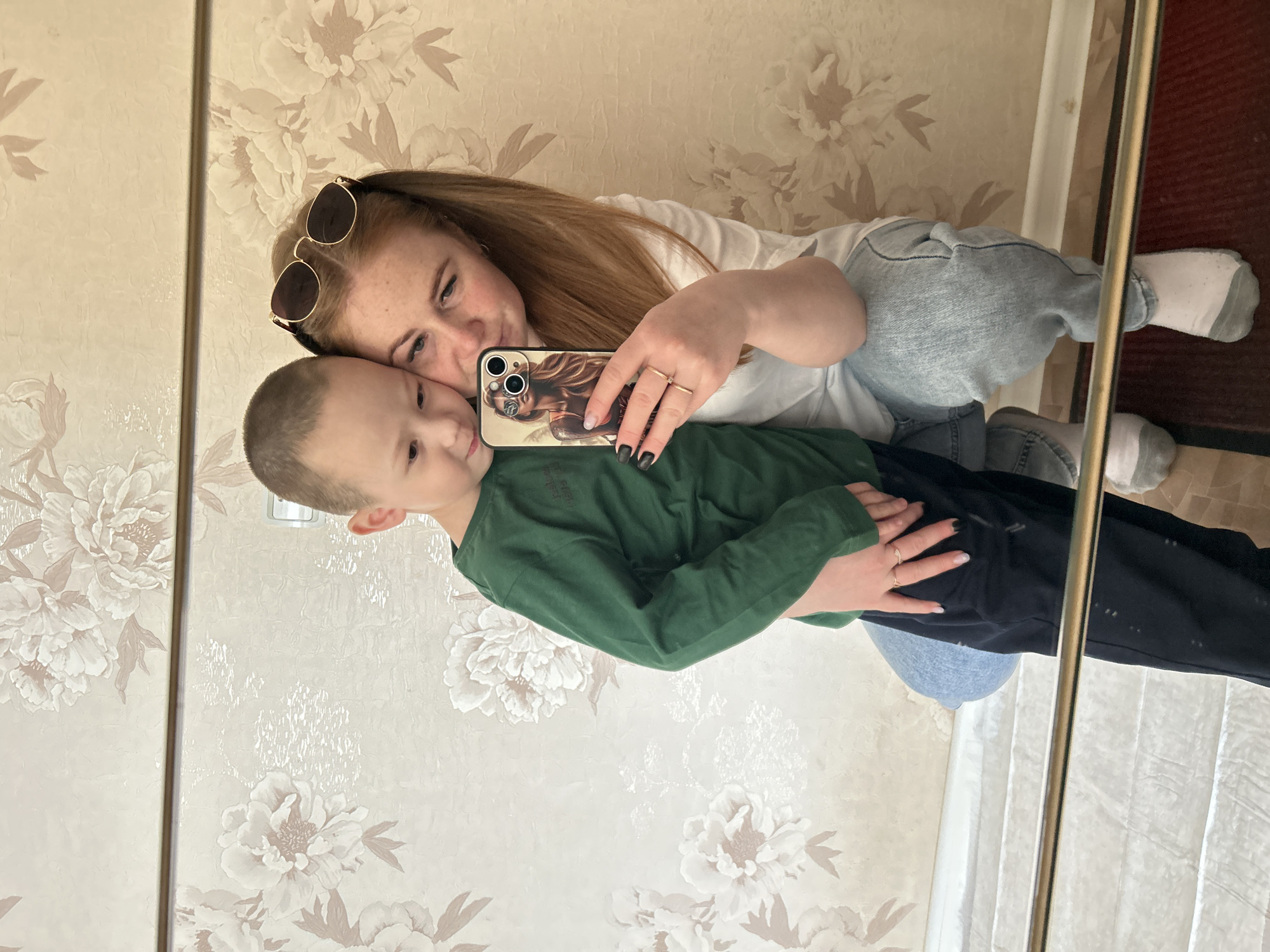 A woman and a young boy are captured in a mirror selfie, taken indoors in Chaikovsky, Russia. The image is oriented vertically, suggesting a 90-degree clockwise rotation from its original capture.
The woman, positioned behind and to the left of the boy, has long, reddish-brown hair with sunglasses pushed up onto her head. She is wearing a white t-shirt, light blue jeans, and white socks with grey soles. She holds a smartphone in her left hand, which features a distinctive phone case depicting a stylized figure with glasses and a hat. Two rings are visible on her left hand, which also has black nail polish. She looks fondly towards the phone with a subtle smile.
The young boy, likely between 4 and 7 years old, stands in front of the woman, leaning against her. He has short brown hair and is dressed in a dark green long-sleeved shirt and dark pants. His expression is calm and neutral as he looks towards the camera.
The background, visible in the mirror reflection, consists of a wall covered in light beige or cream-colored wallpaper with prominent floral patterns, resembling large peonies. A vertical wooden or metallic strip divides sections of the wall decoration. On the far right side of the reflection, a glimpse of a dark reddish-brown floor or rug and a white curtain or blind is visible. The indoor lighting suggests it is daytime. No discernible text is visible within the image.